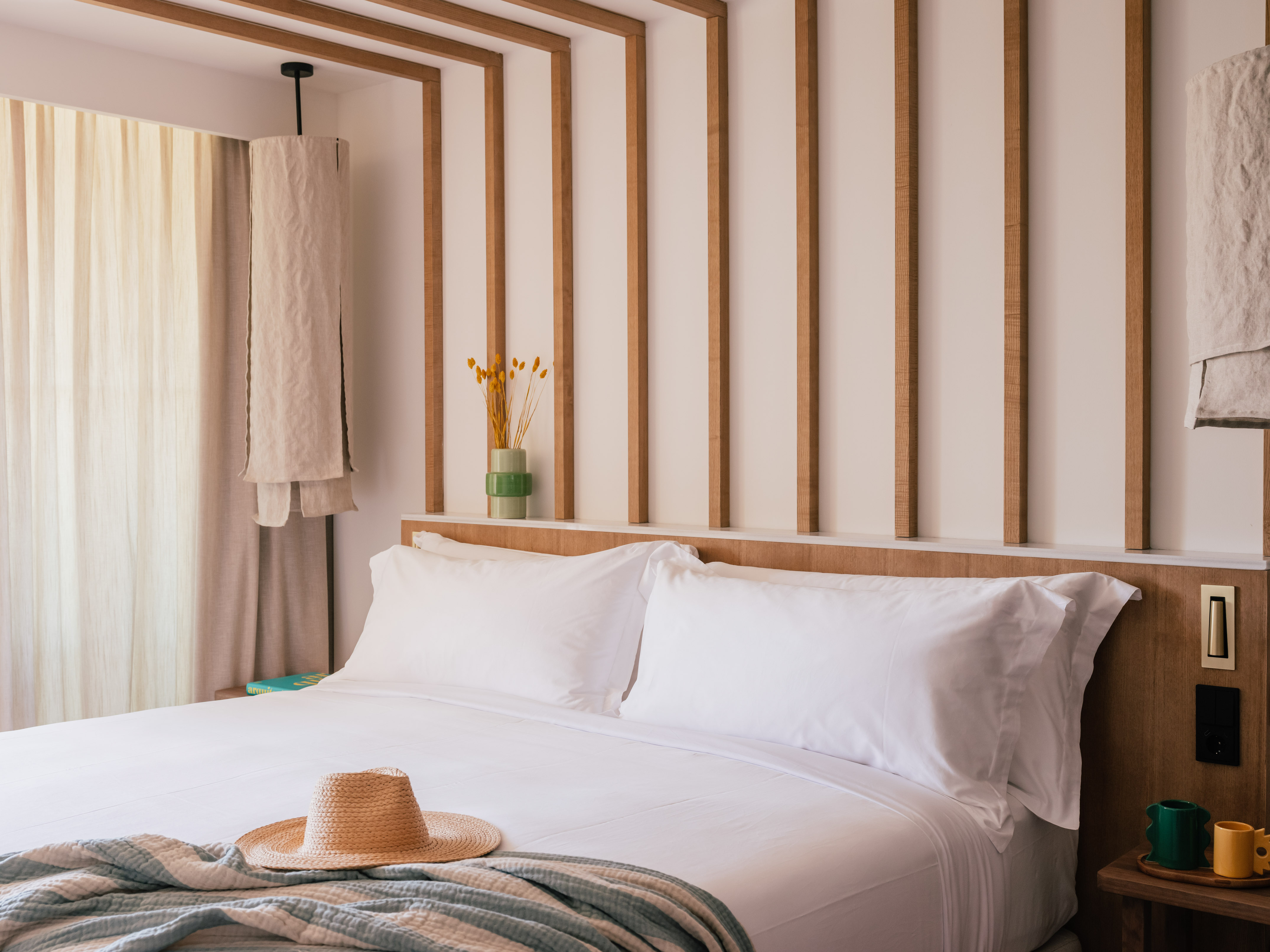 Bright hotel room with white bed, straw hat, and wooden slat headboard.