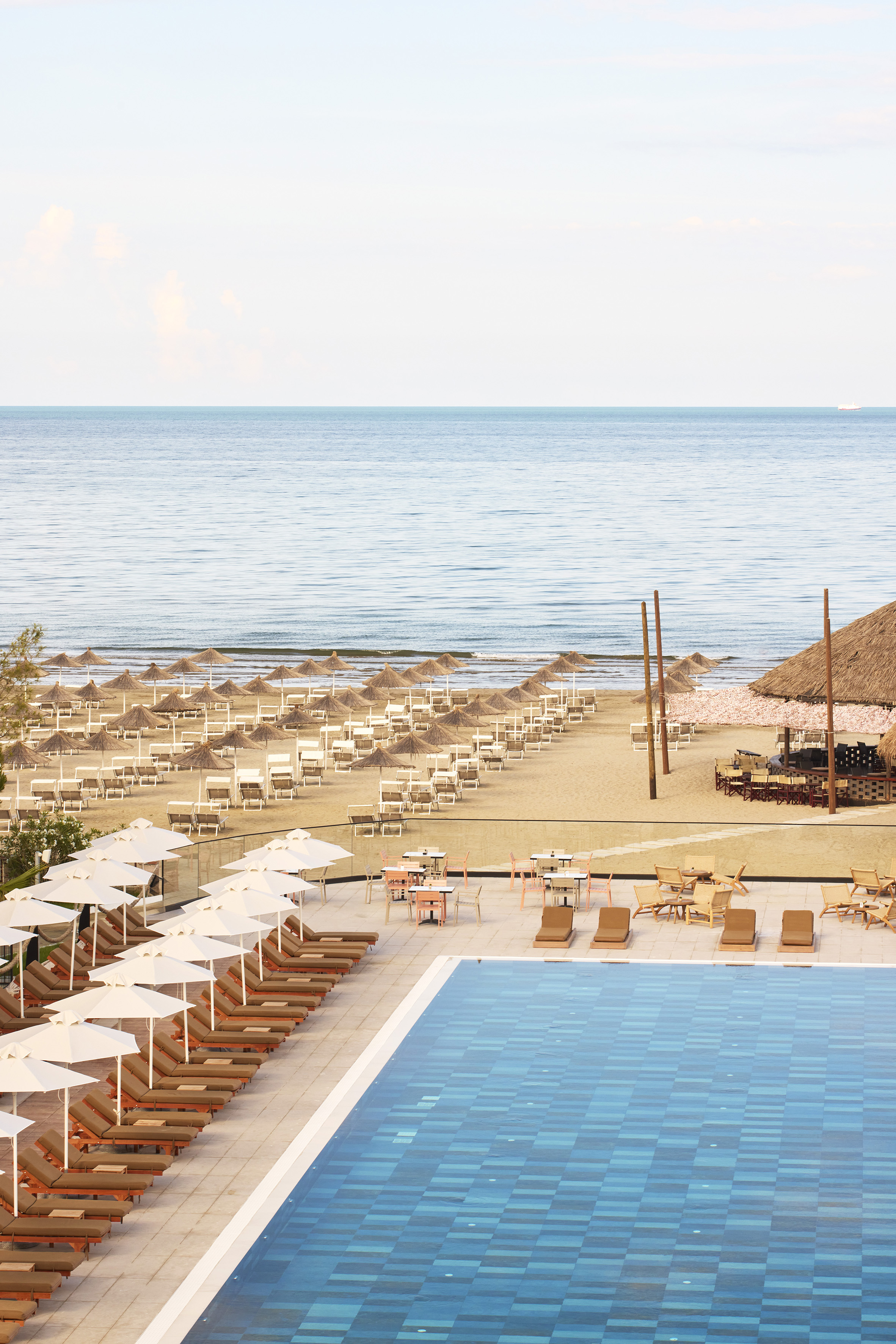 Resort swimming pool, sandy beach with sun loungers, straw umbrellas, and calm ocean.