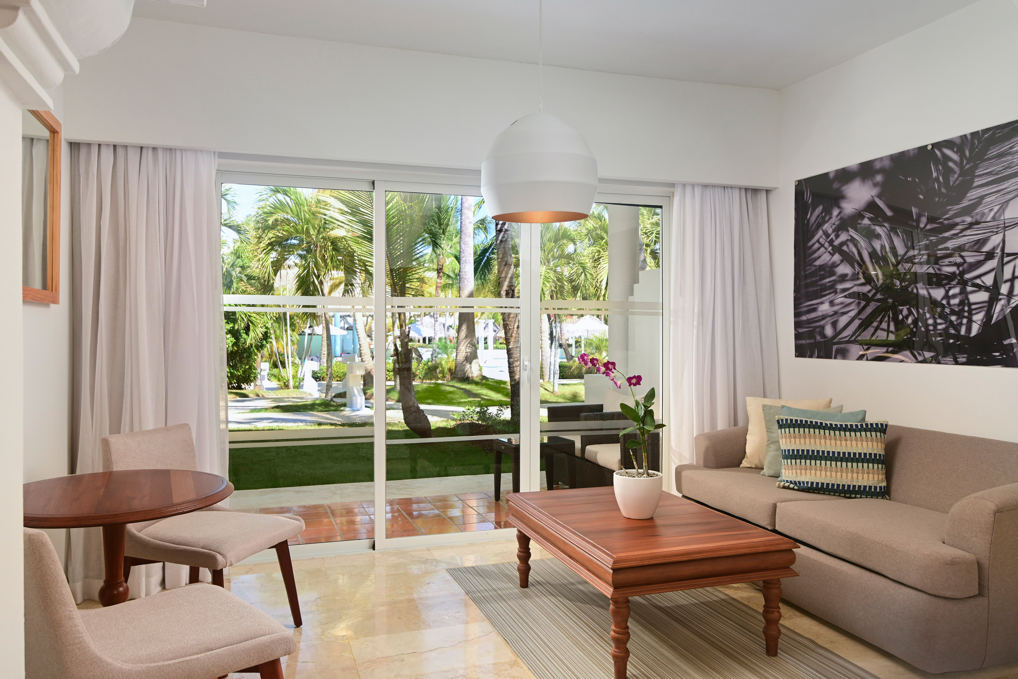 Spacious hotel living room with sofa, coffee table, and tropical palm garden view.