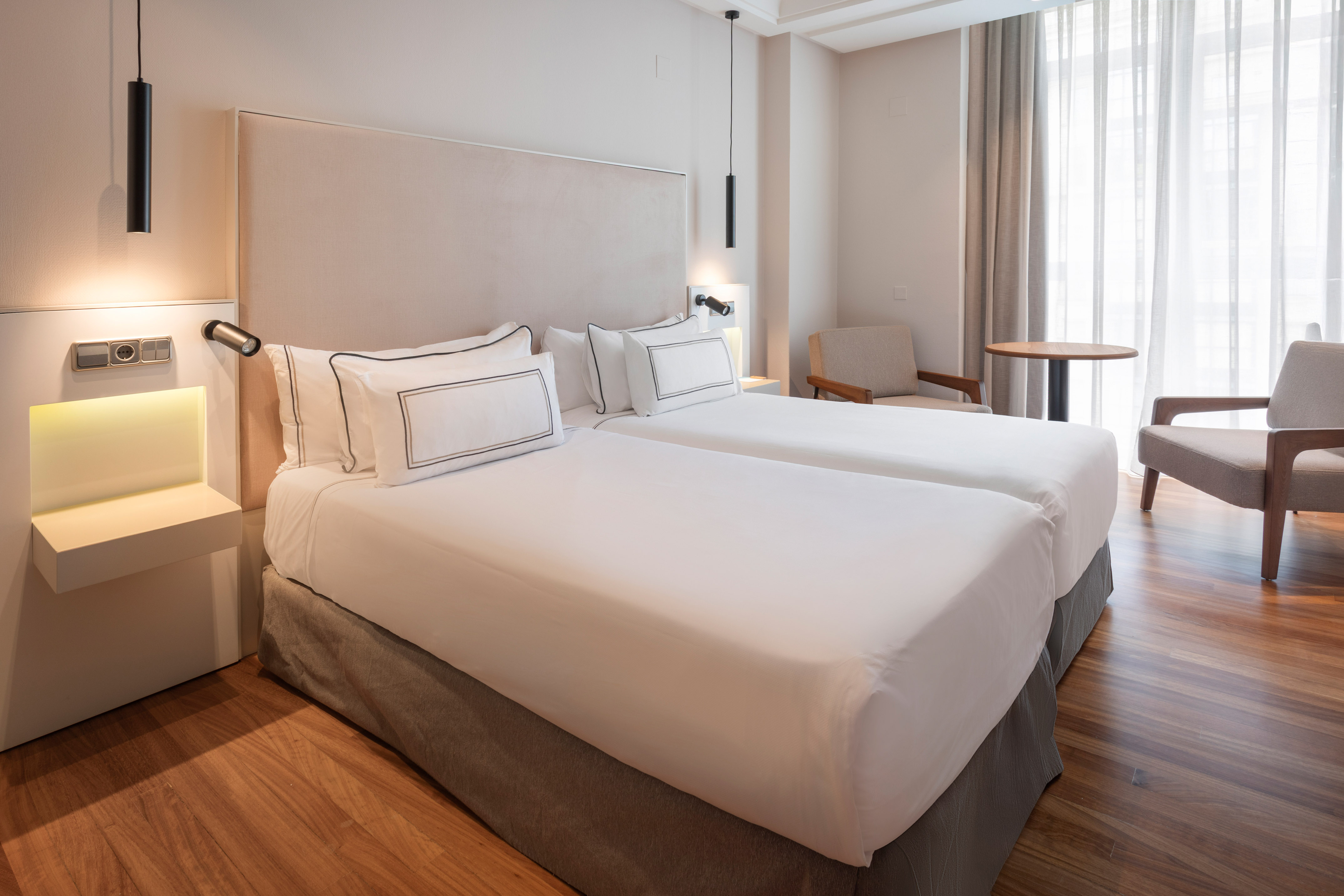 Modern hotel room with twin beds, hardwood floor, neutral decor, and seating area.
