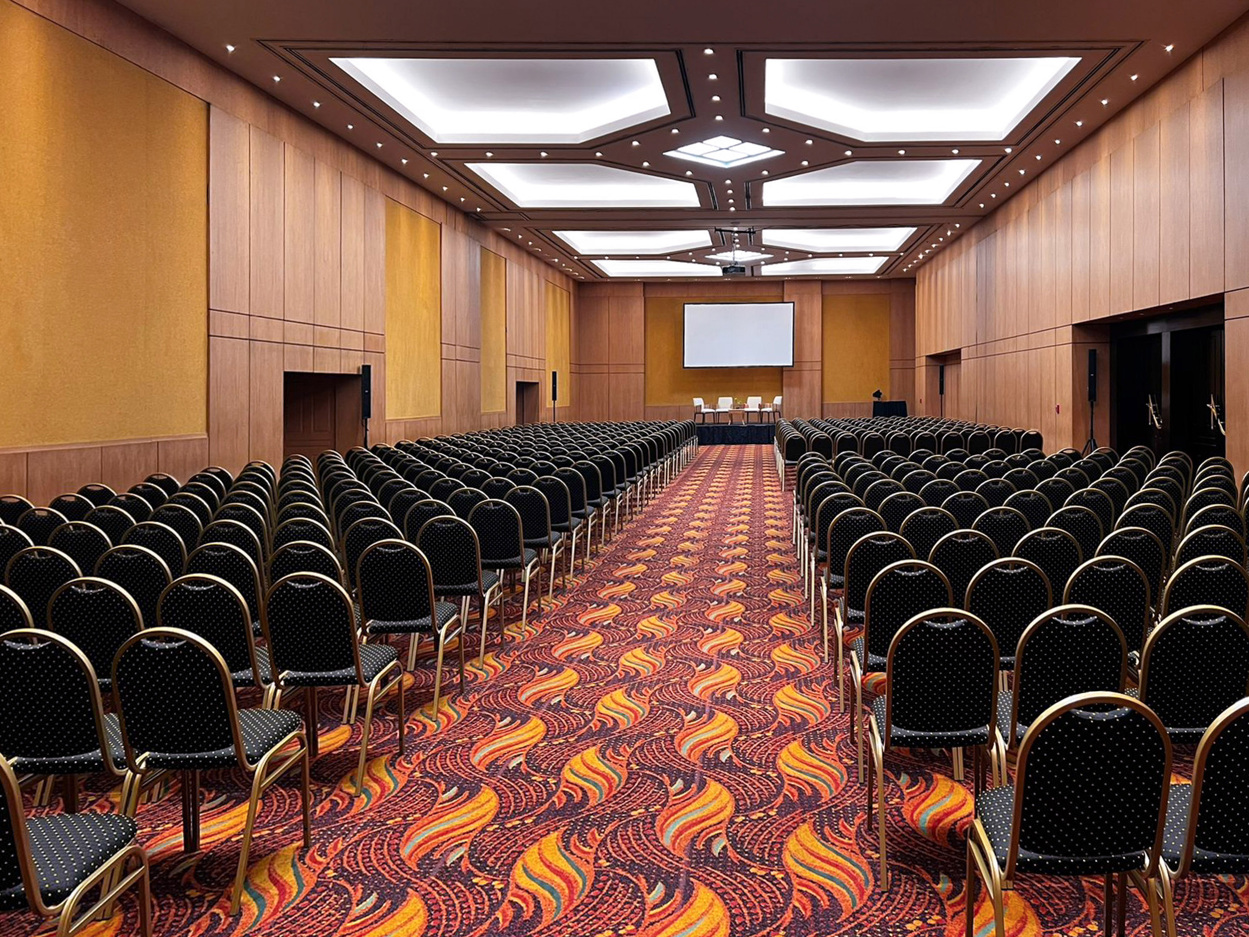 Large conference hall with rows of black chairs, projection screen, and patterned carpet.