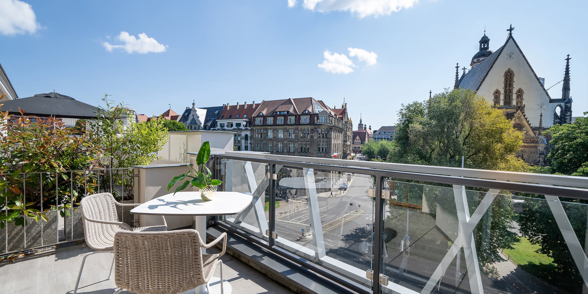 Rooftop balcony with glass railing, outdoor seating, and Leipzig city view with church.