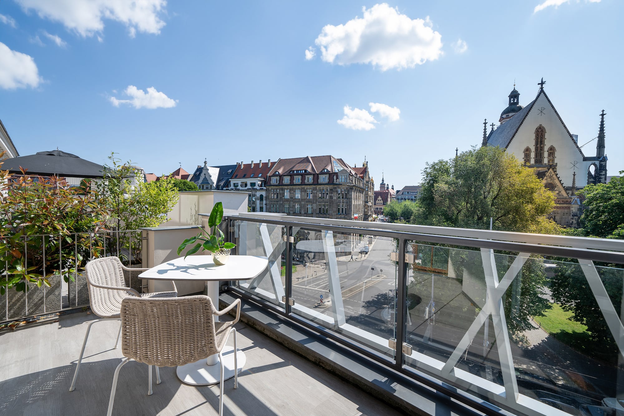 Rooftop balcony with glass railing, outdoor seating, and Leipzig city view with church.