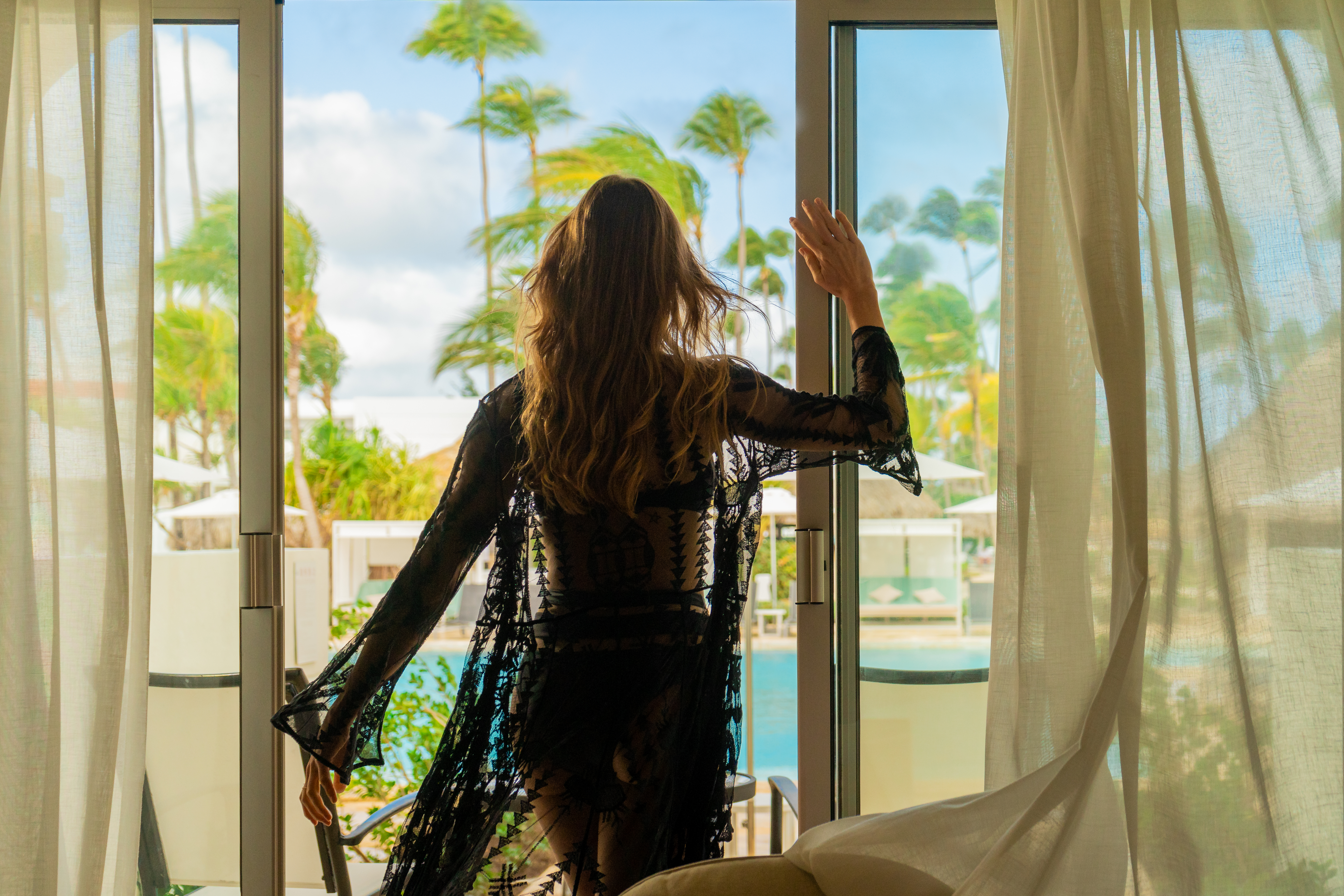 Woman in black sheer robe looks out at tropical resort, palm trees, pool.