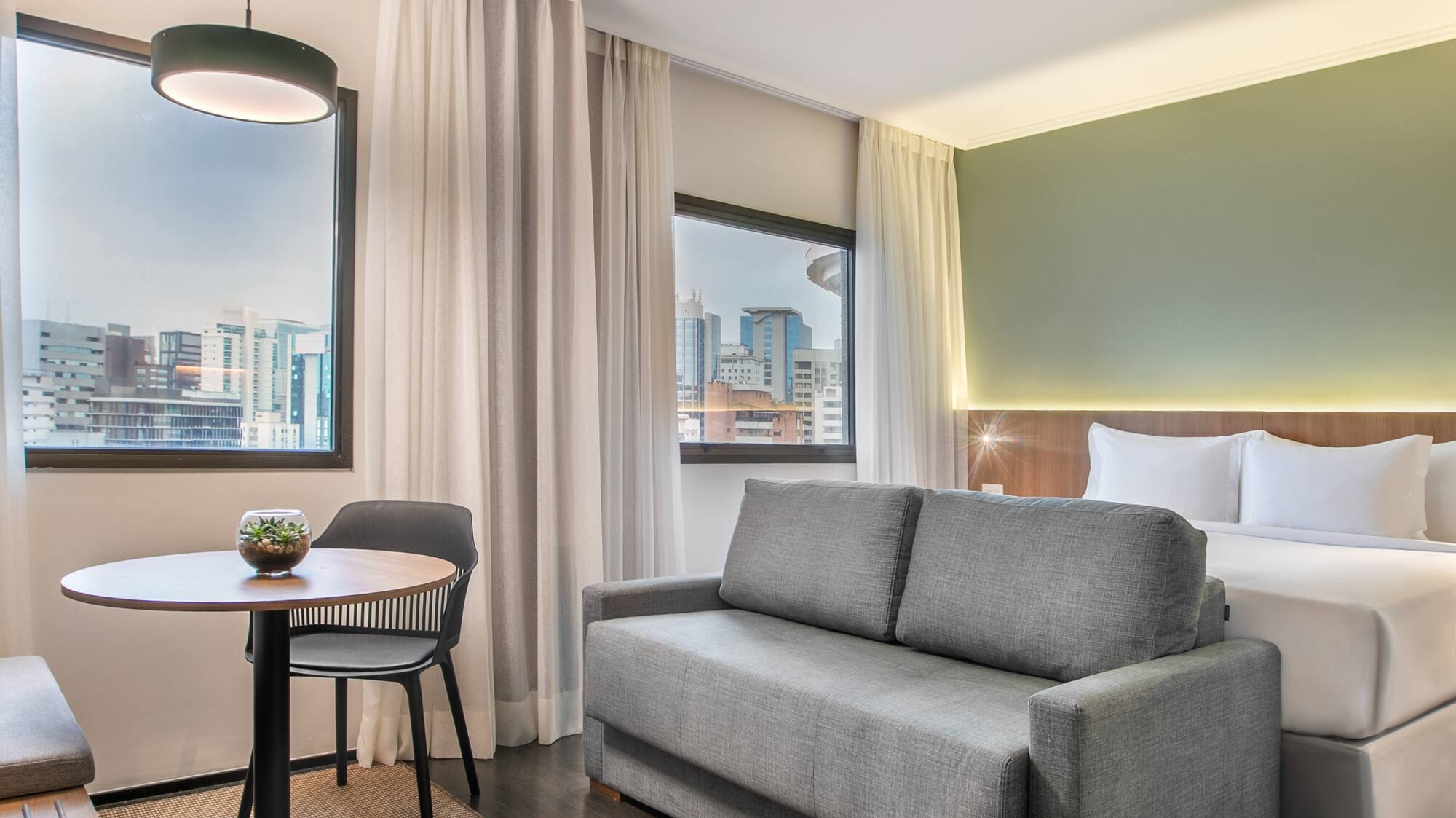 Modern hotel suite with bed, grey sofa, dining area, and city view.