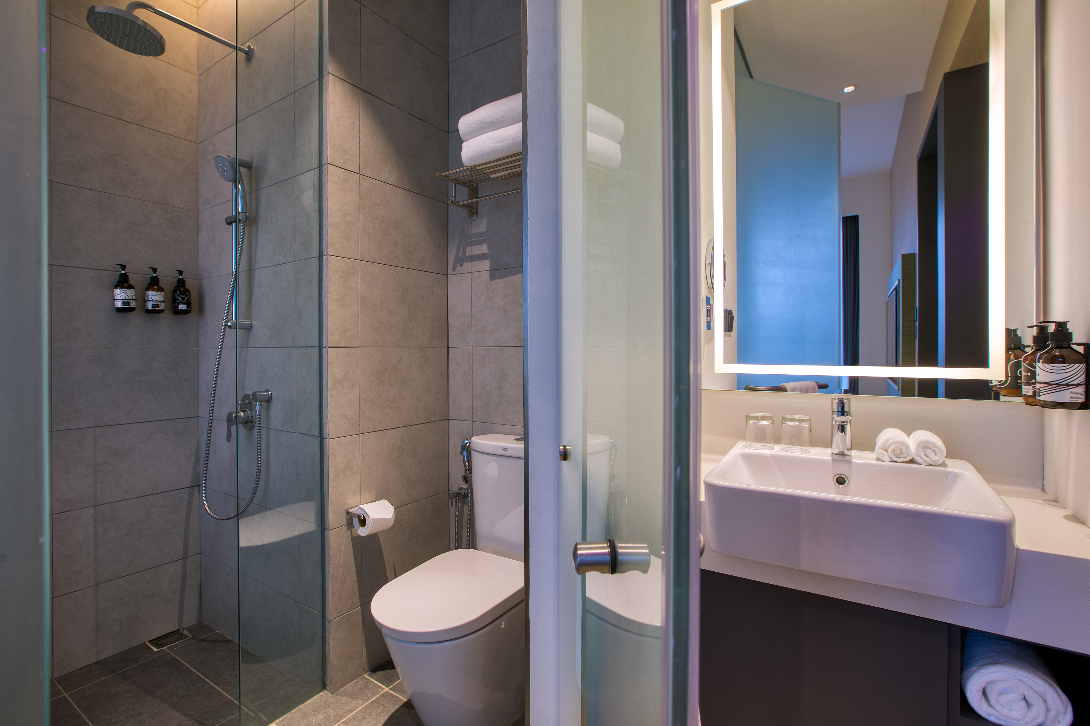 Modern tiled bathroom with glass shower, toilet, vanity, and illuminated mirror.