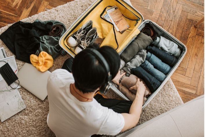 Man with headphones packing travel suitcase with clothes, laptop, and mask.