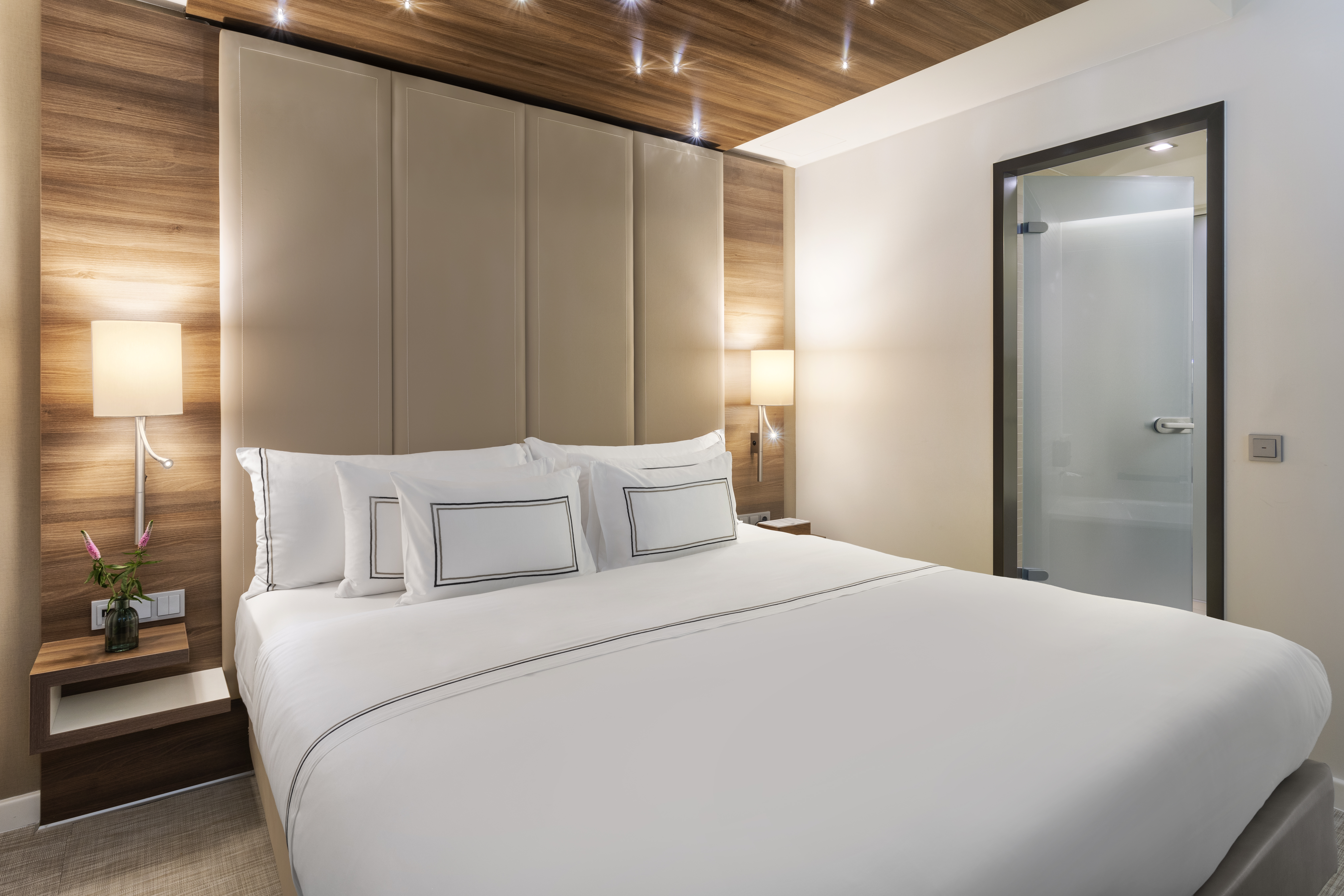 Modern hotel room with white bed, padded headboard, wood accents, and lamps.