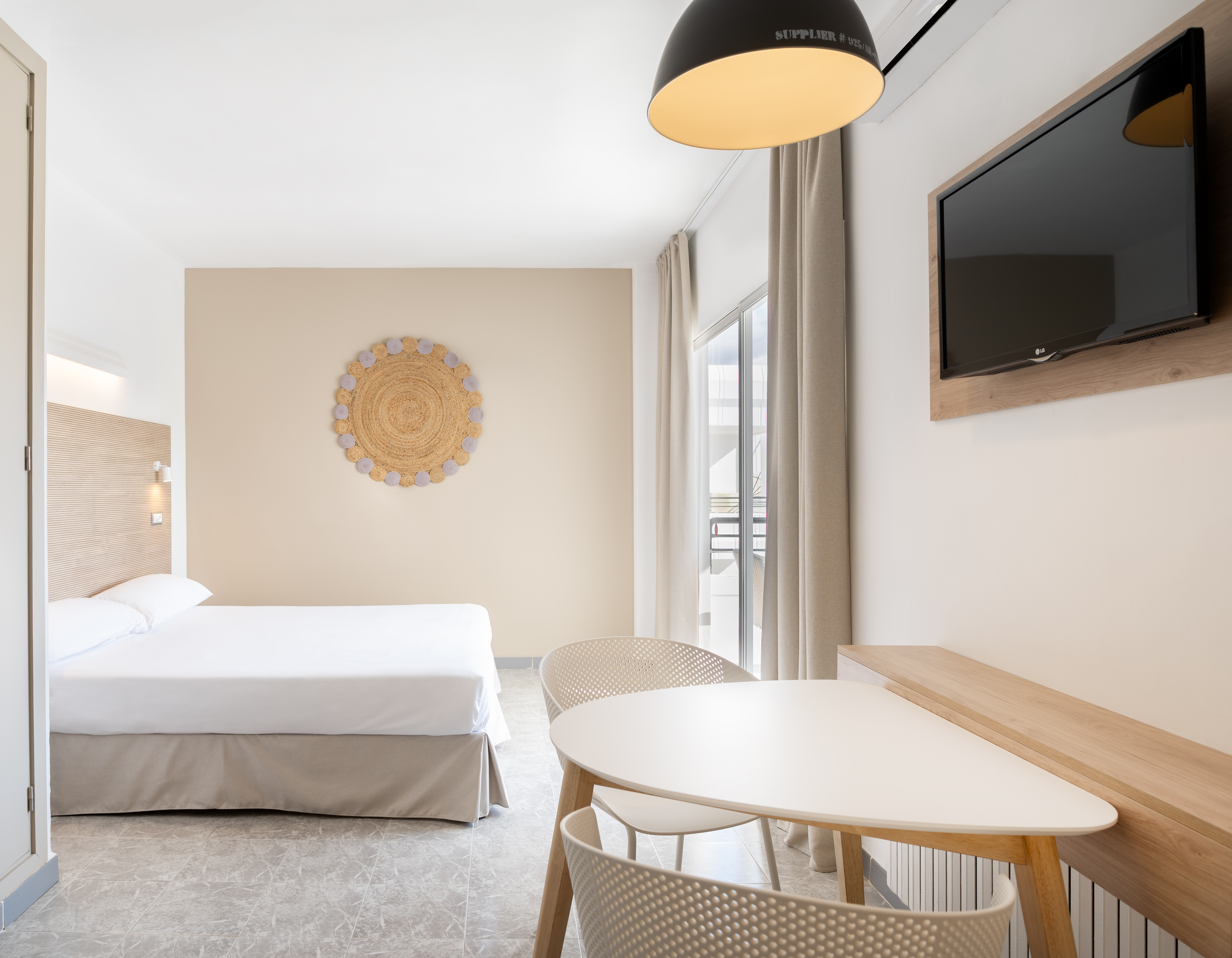 Bright modern hotel room with bed, dining table, TV, and woven wall art.