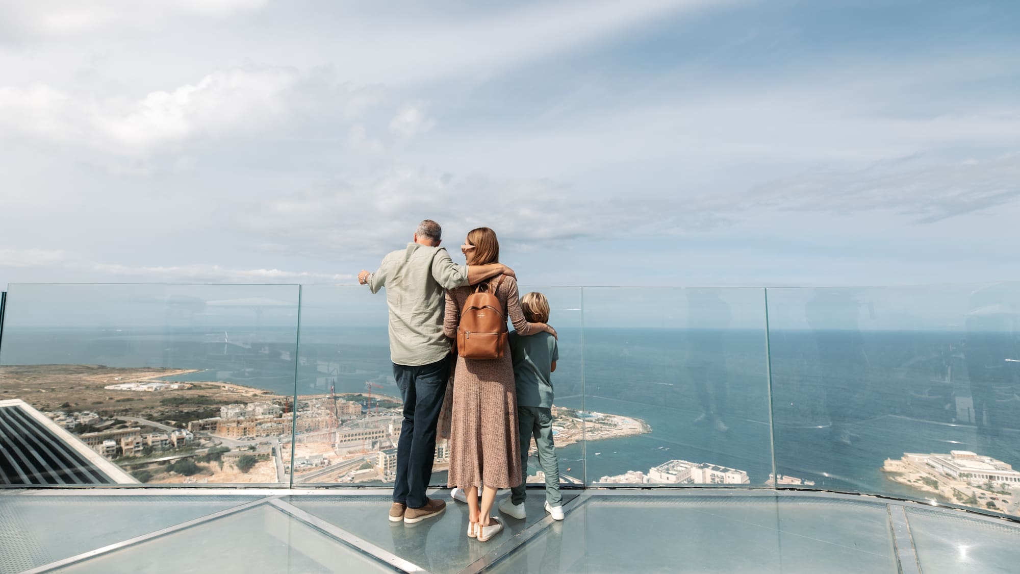 Family on glass skywalk overlooking panoramic ocean and cityscape.