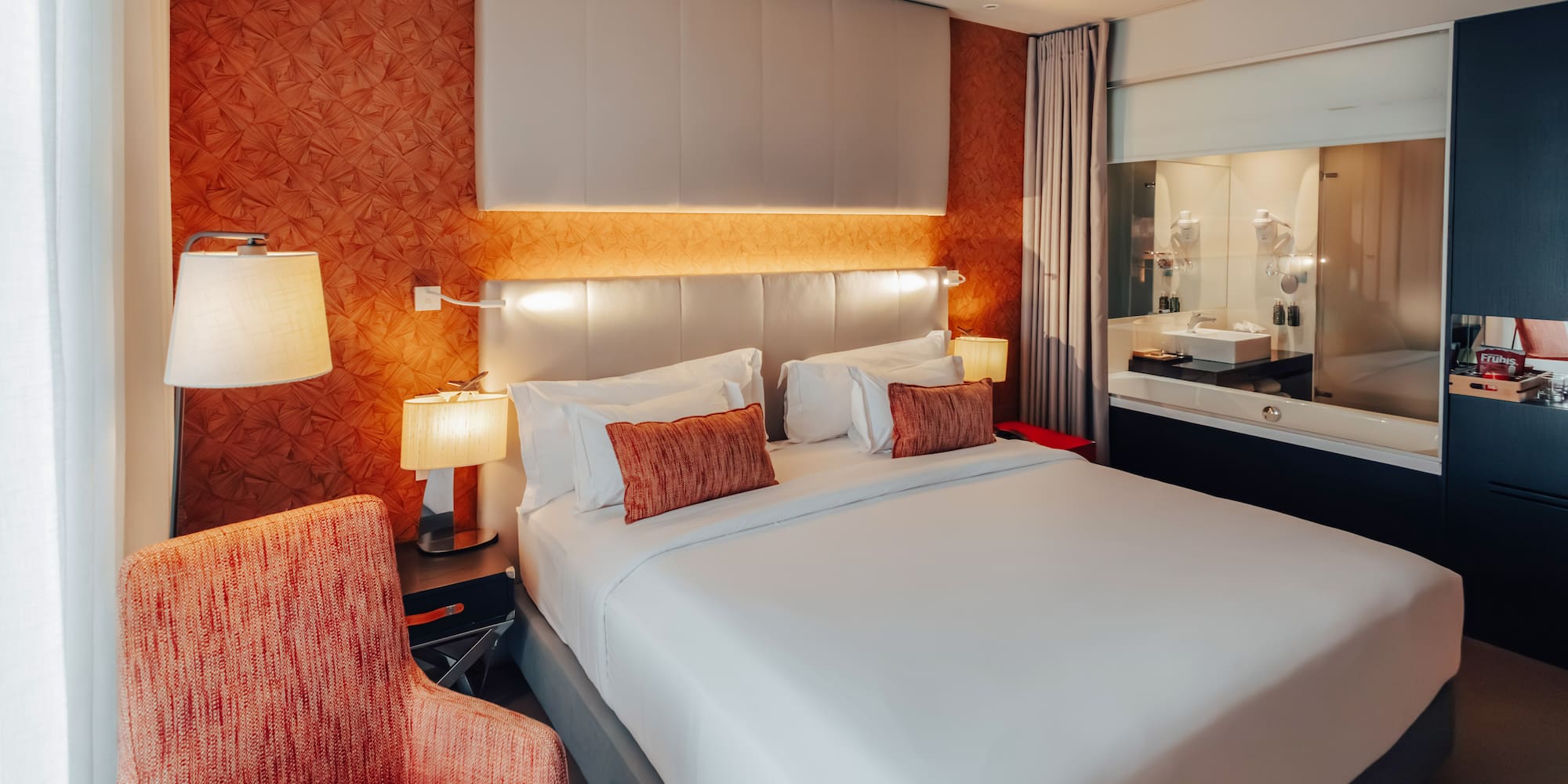 Modern hotel bedroom with king bed, orange accent wall, and integrated bathroom.