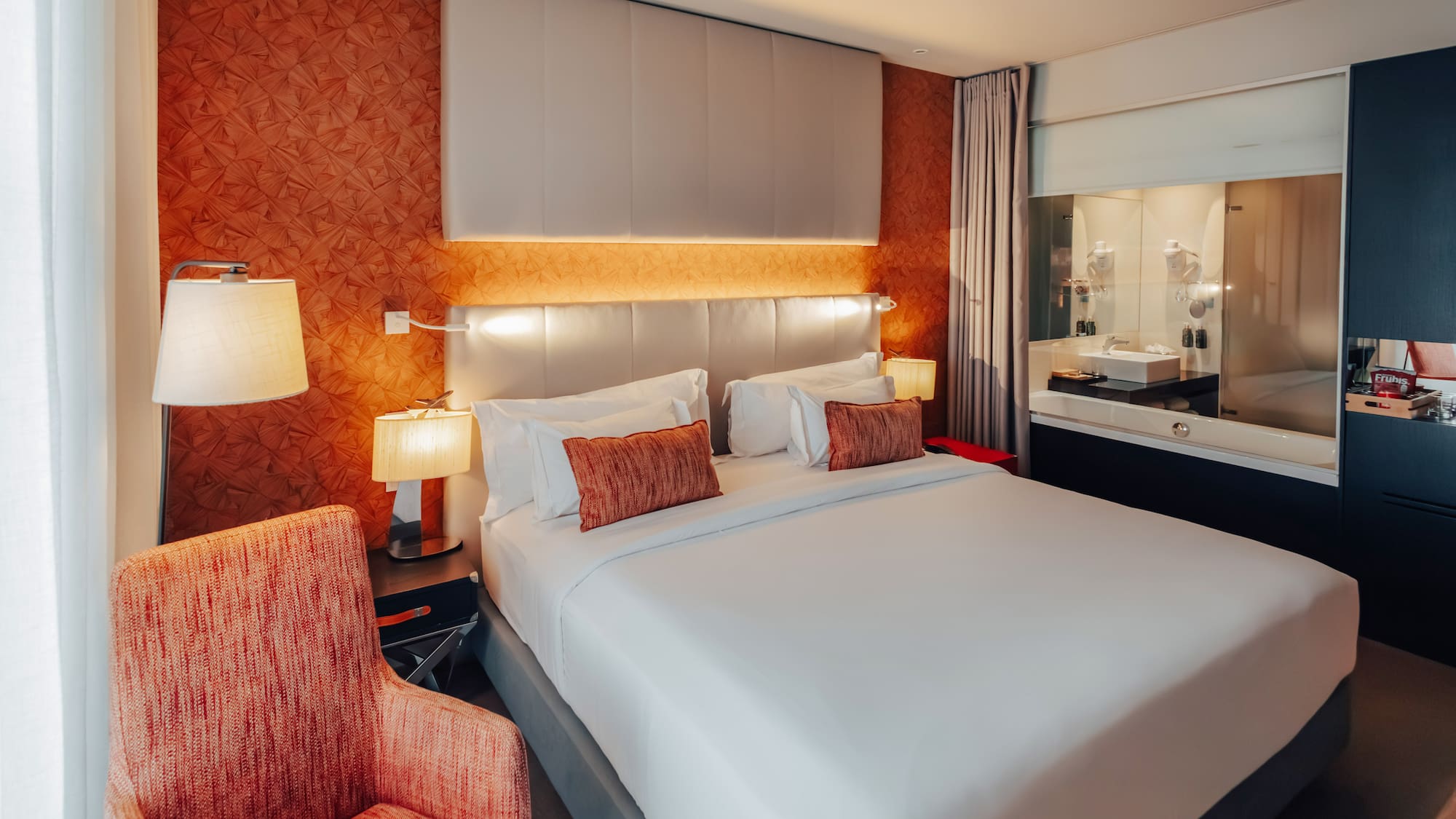 Modern hotel bedroom with king bed, orange accent wall, and integrated bathroom.
