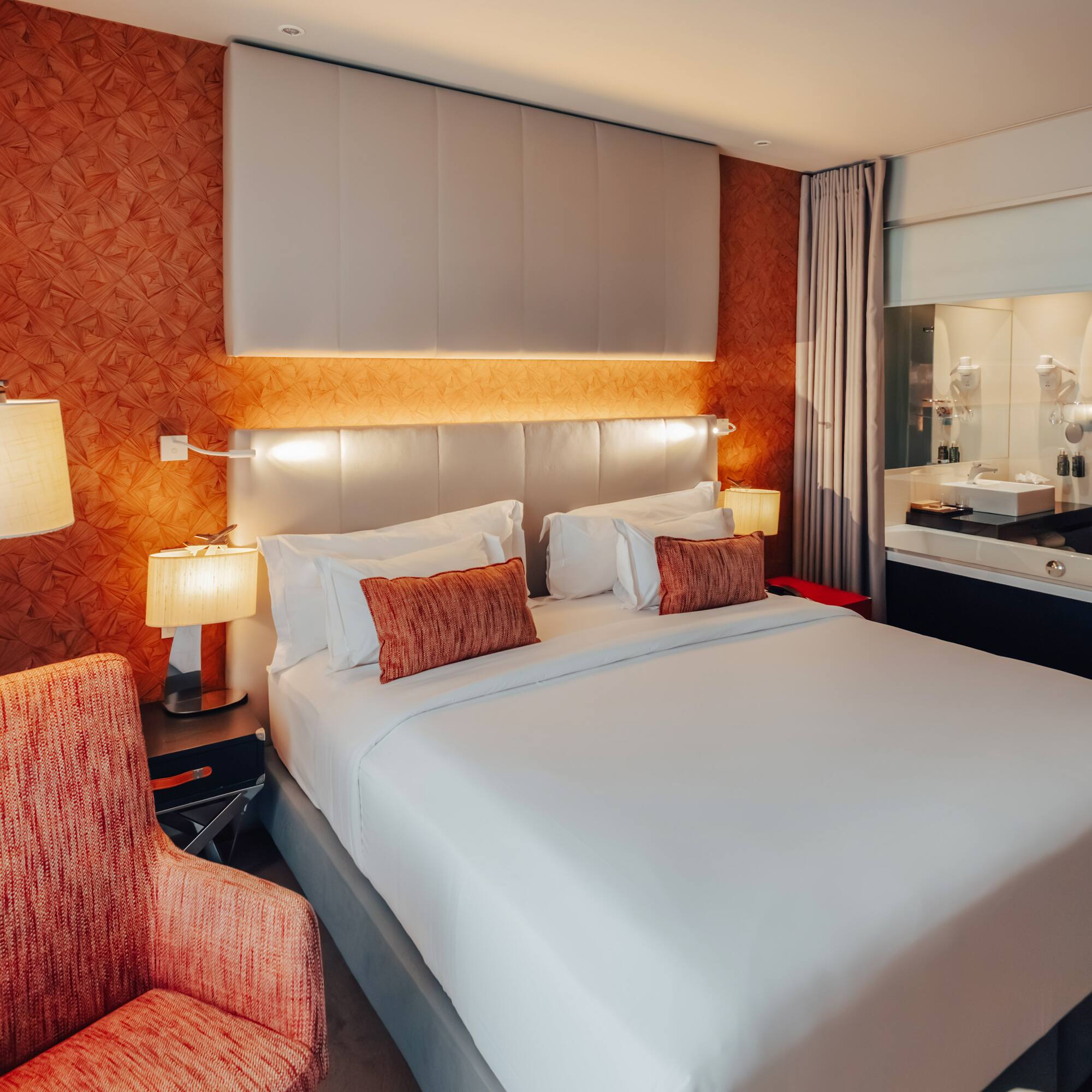 Modern hotel bedroom with king bed, orange accent wall, and integrated bathroom.