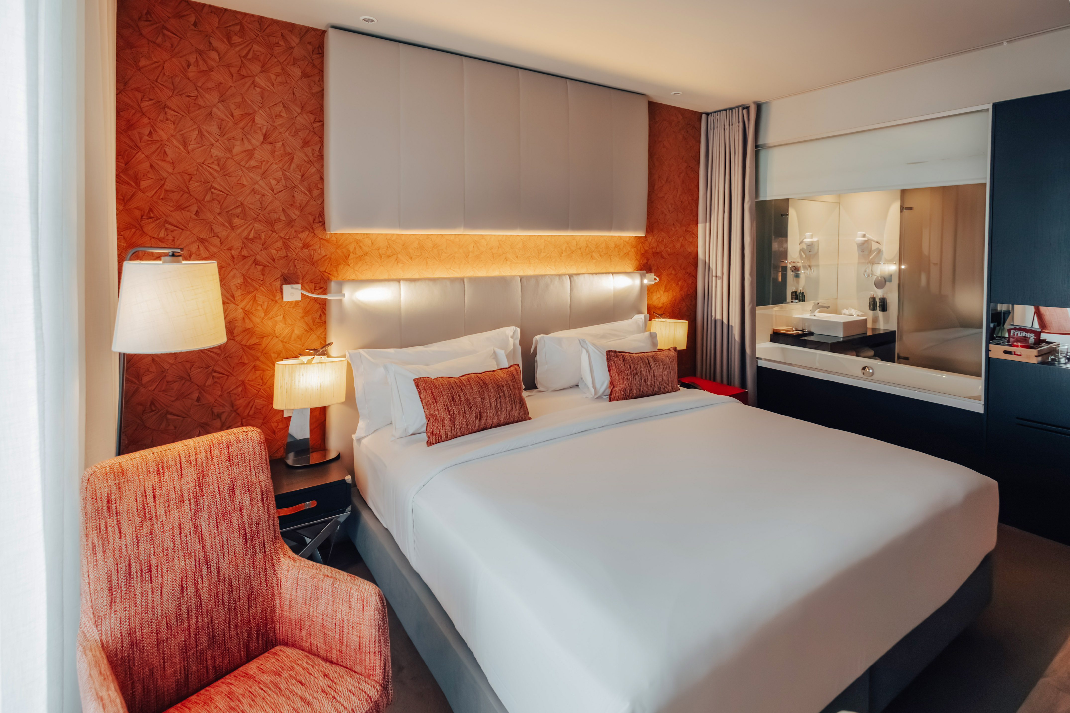 Modern hotel bedroom with king bed, orange accent wall, and integrated bathroom.