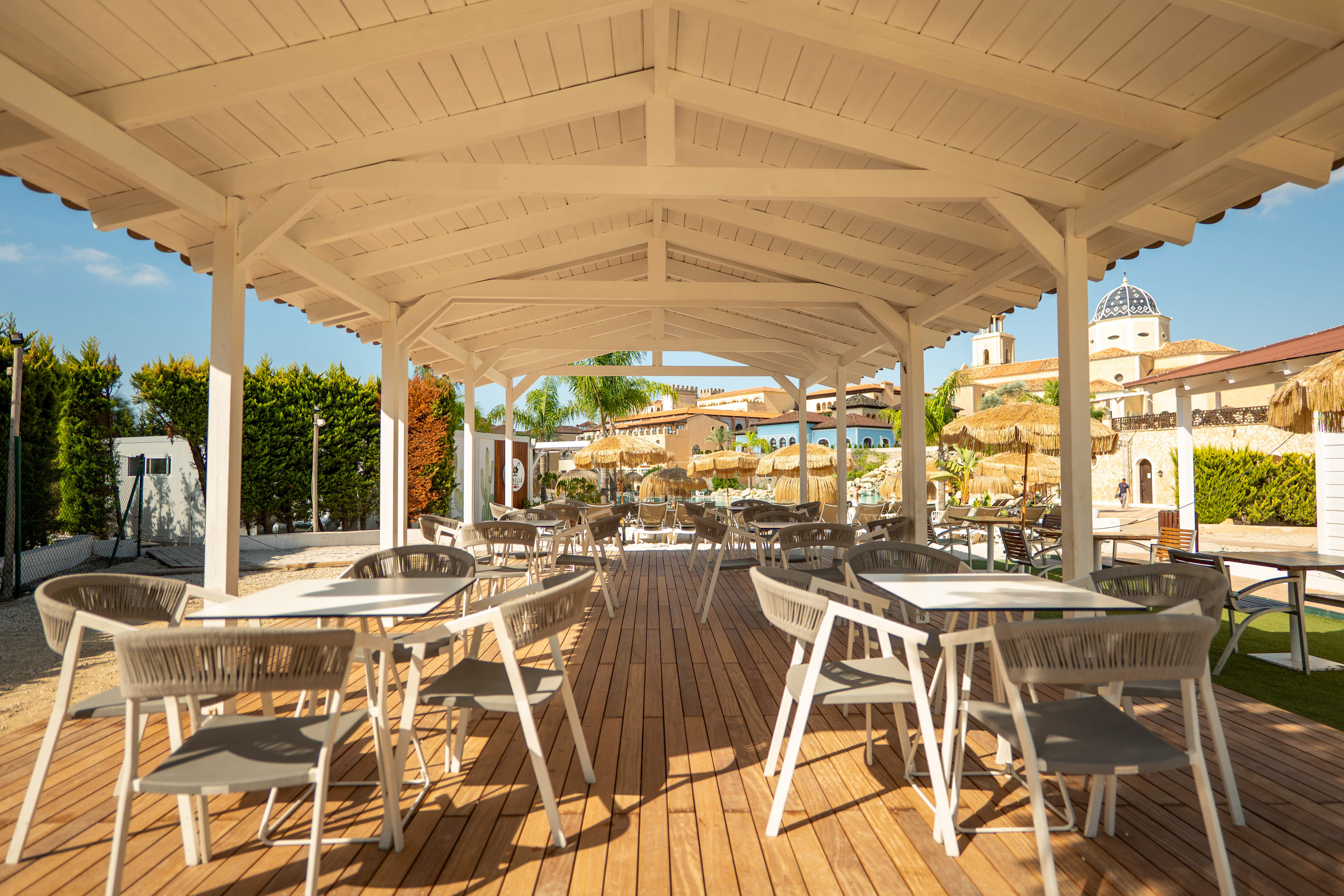 Outdoor restaurant patio under white pergola with wooden deck, resort background.