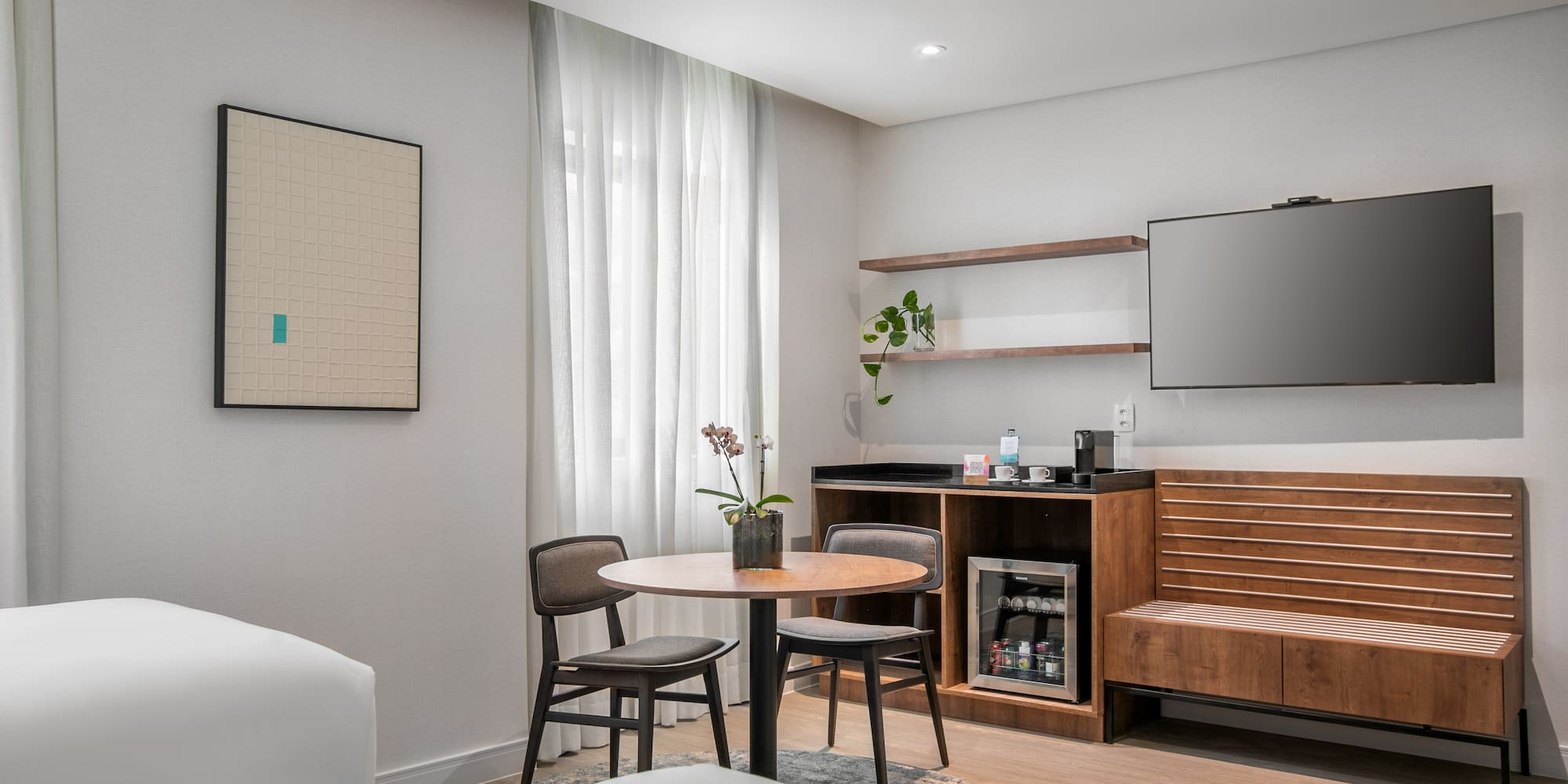 Bright hotel room interior with TV, minibar, round dining table, and art.