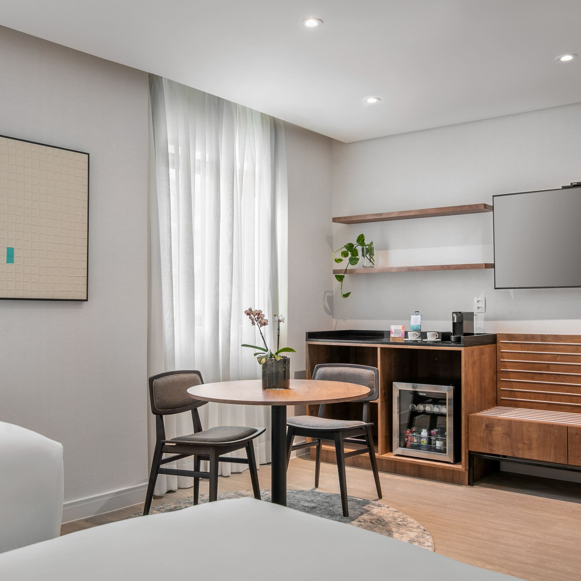 Bright hotel room interior with TV, minibar, round dining table, and art.