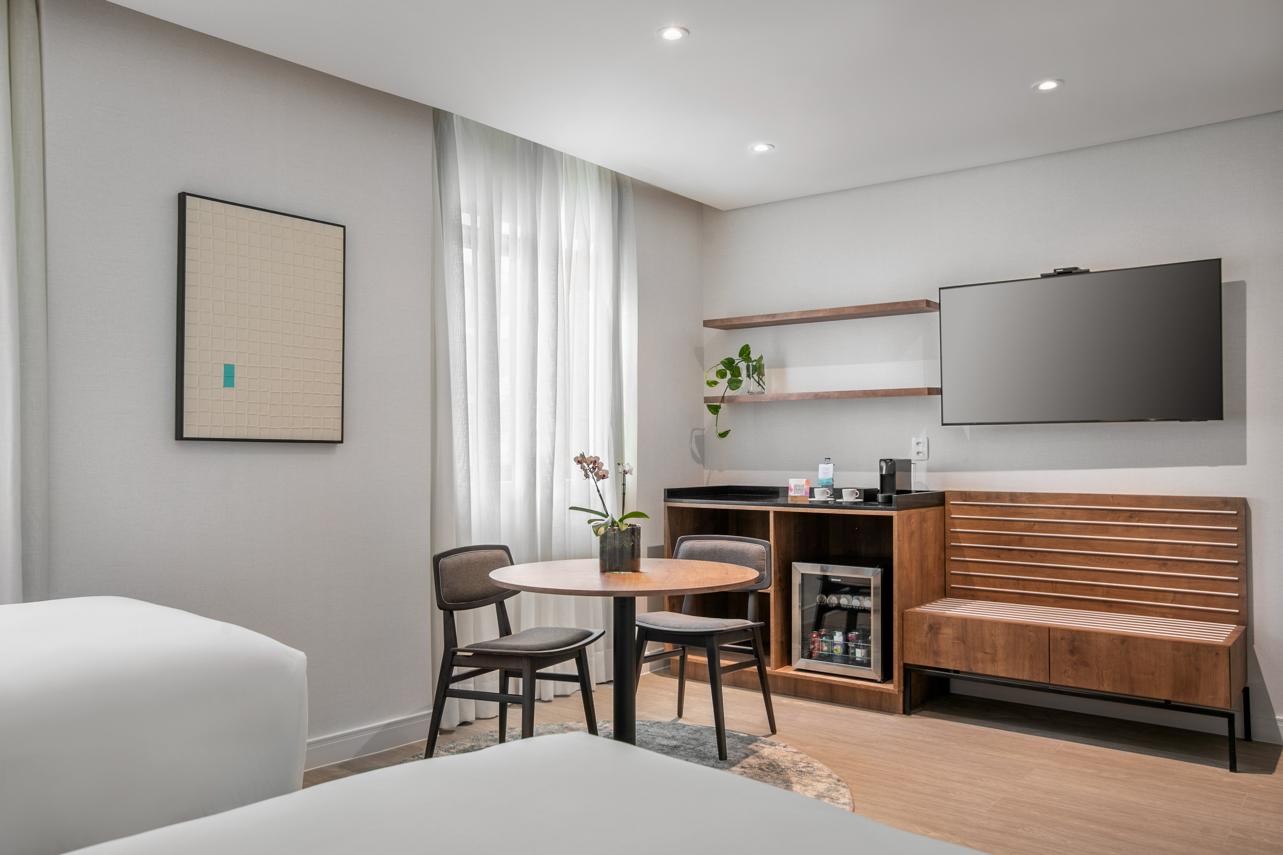 Bright hotel room interior with TV, minibar, round dining table, and art.