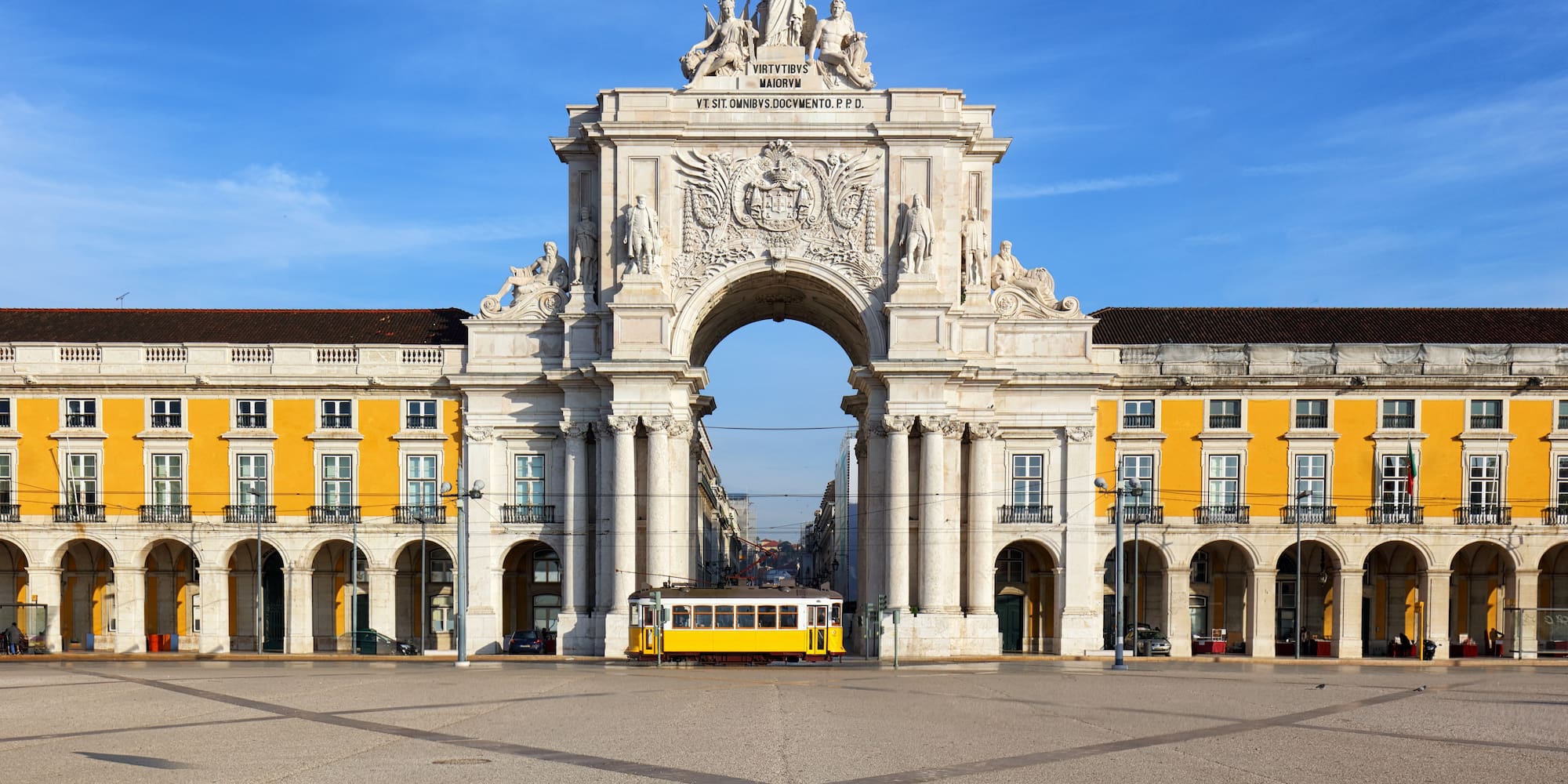 a large white archway with statues and a yellow trolley in front of it with Praça do Comércio in the background