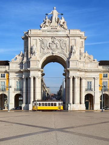a large white archway with statues and a yellow trolley in front of it with Praça do Comércio in the background
