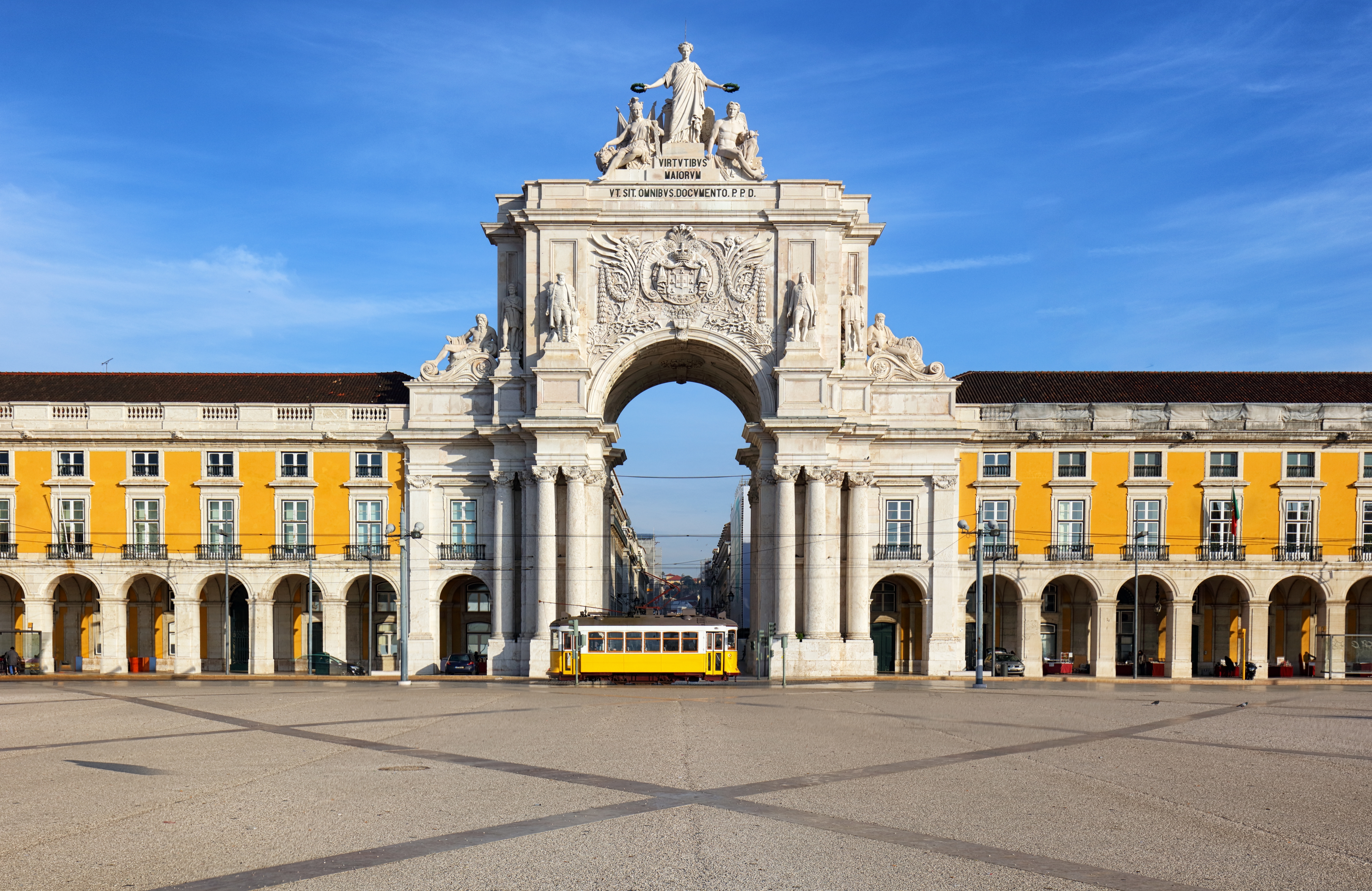 a large white archway with statues and a yellow trolley in front of it with Praça do Comércio in the background