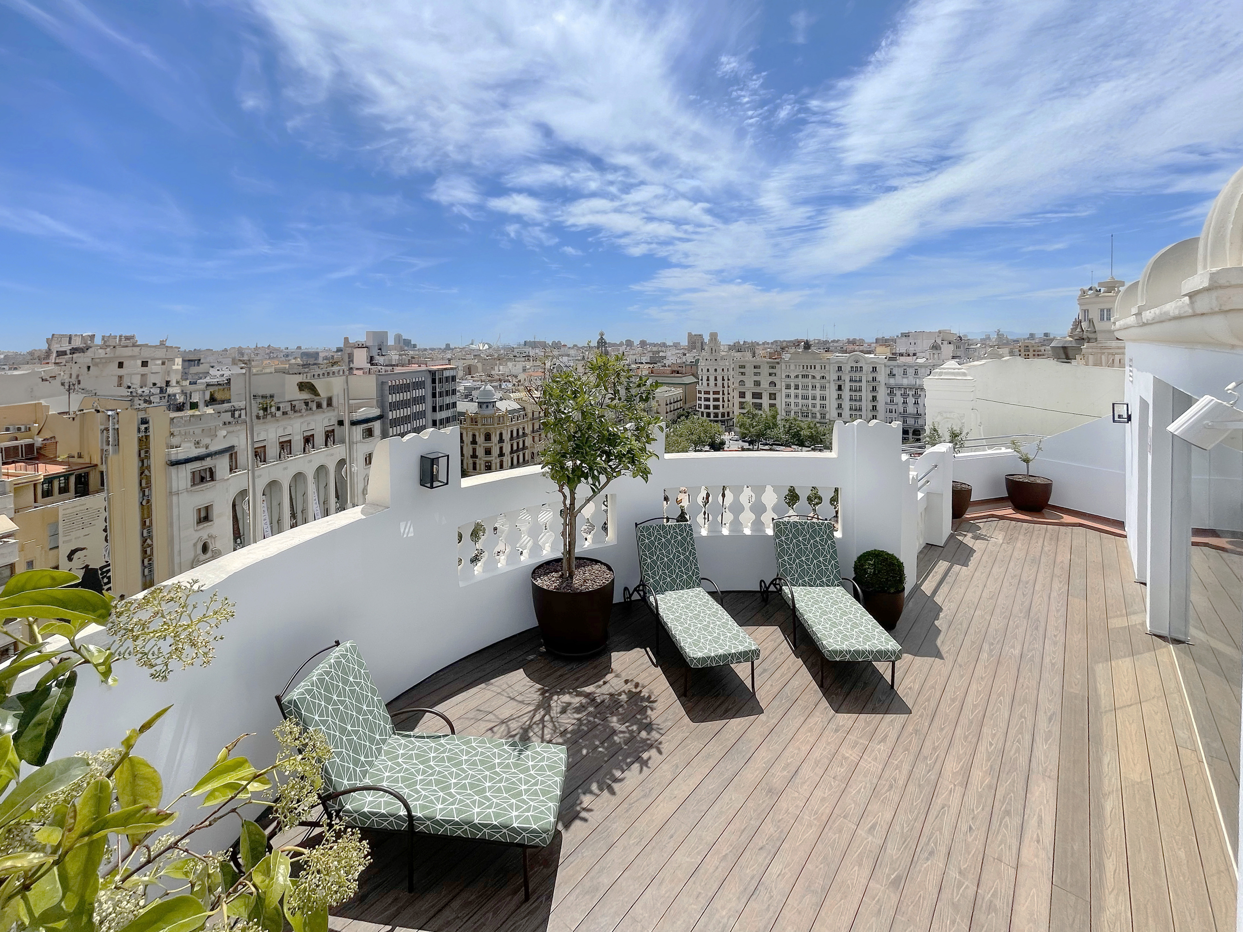 Sunny rooftop terrace with green lounge chairs overlooking city skyline.