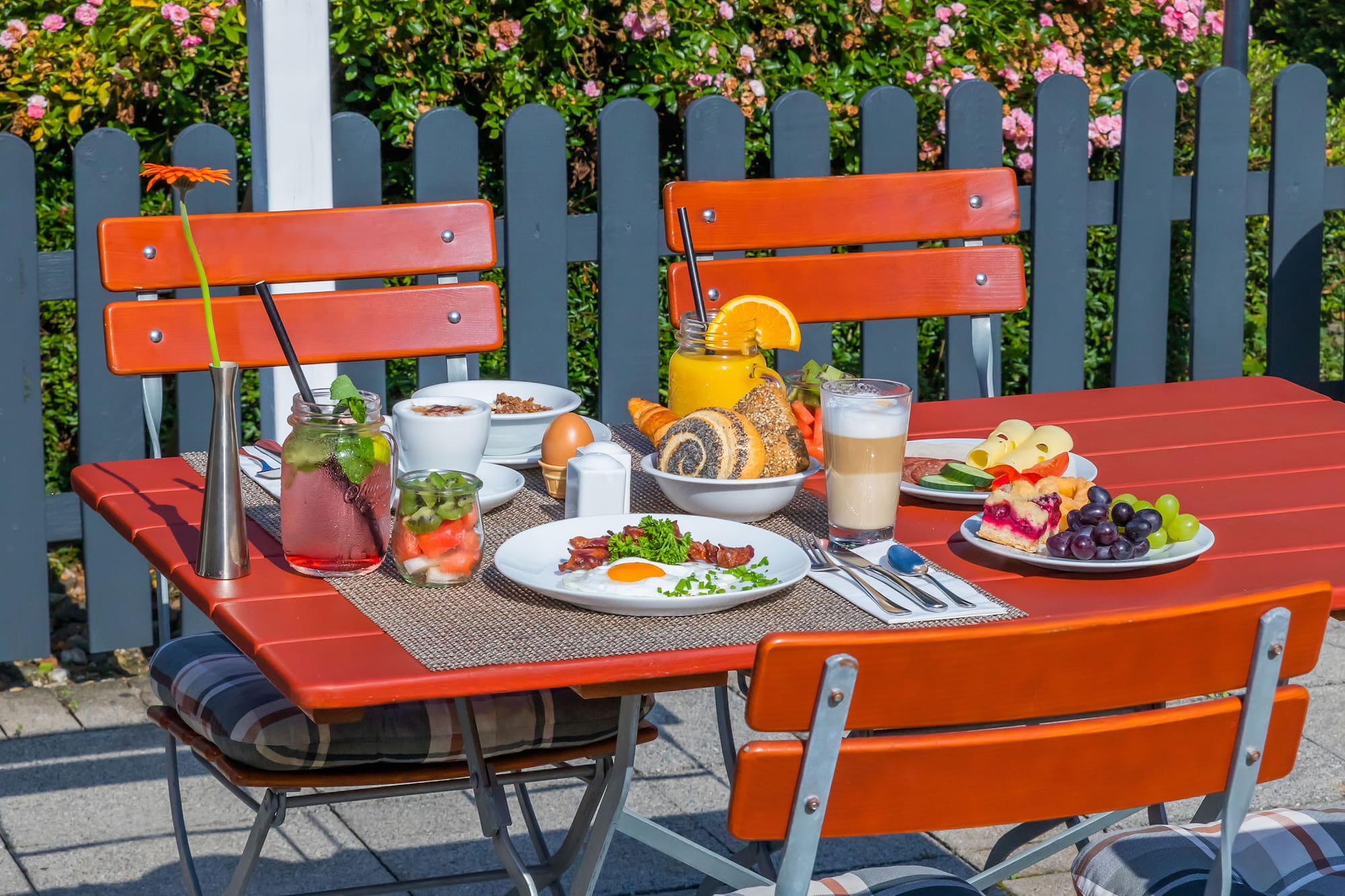 Outdoor brunch on red patio table with diverse foods and drinks, pink roses.