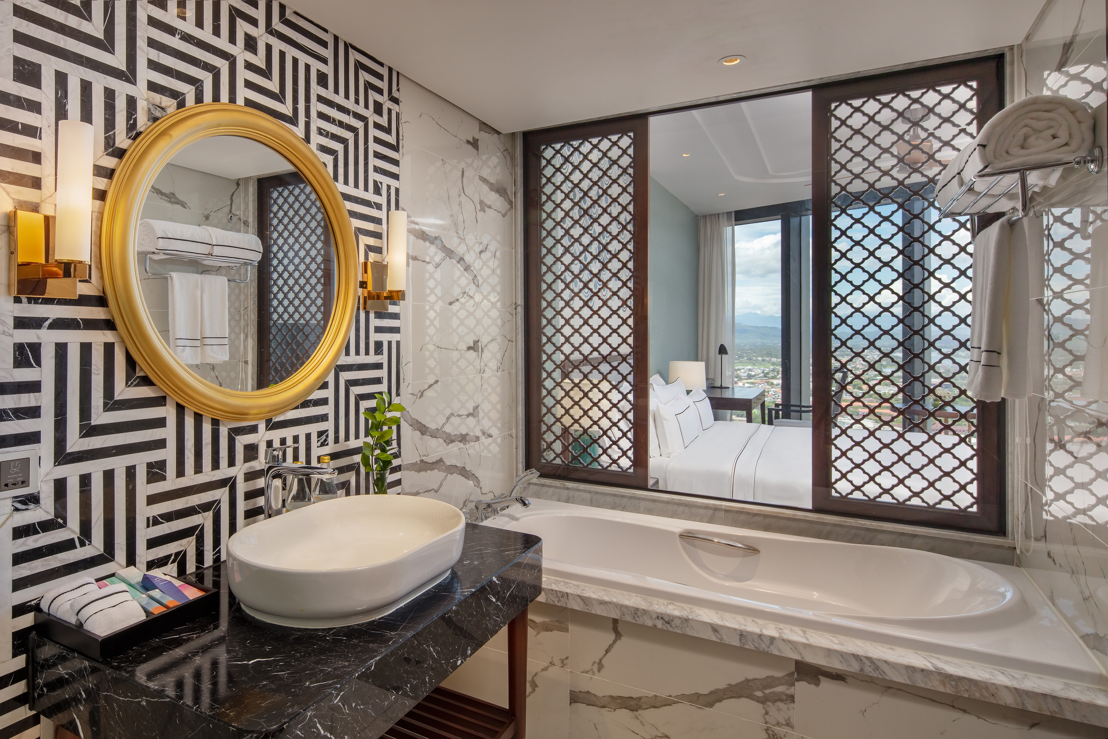 Modern luxury bathroom with geometric tiles, marble, bathtub, and bedroom view.