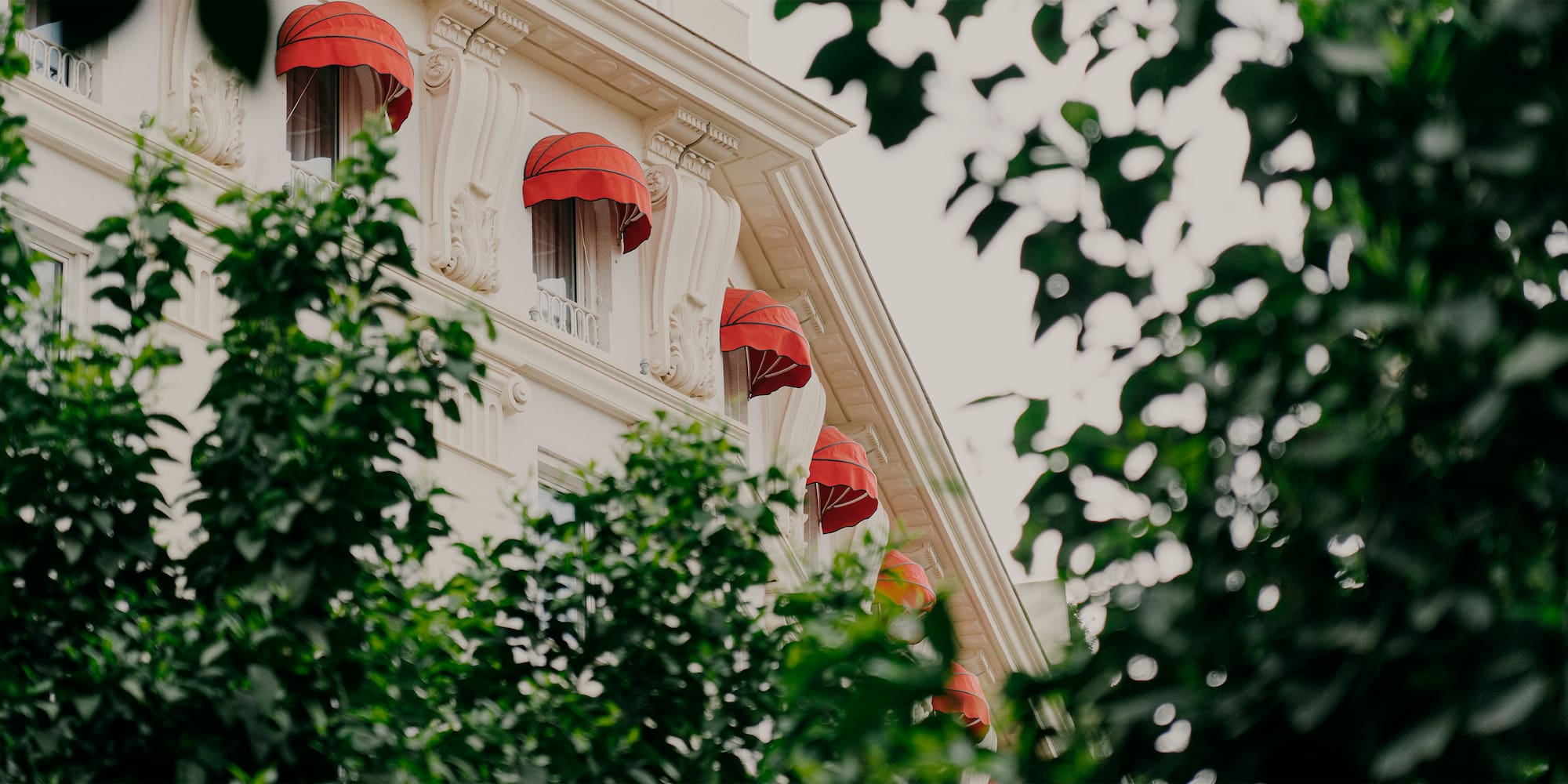 Classic white building facade with red awnings on windows, framed by green trees.