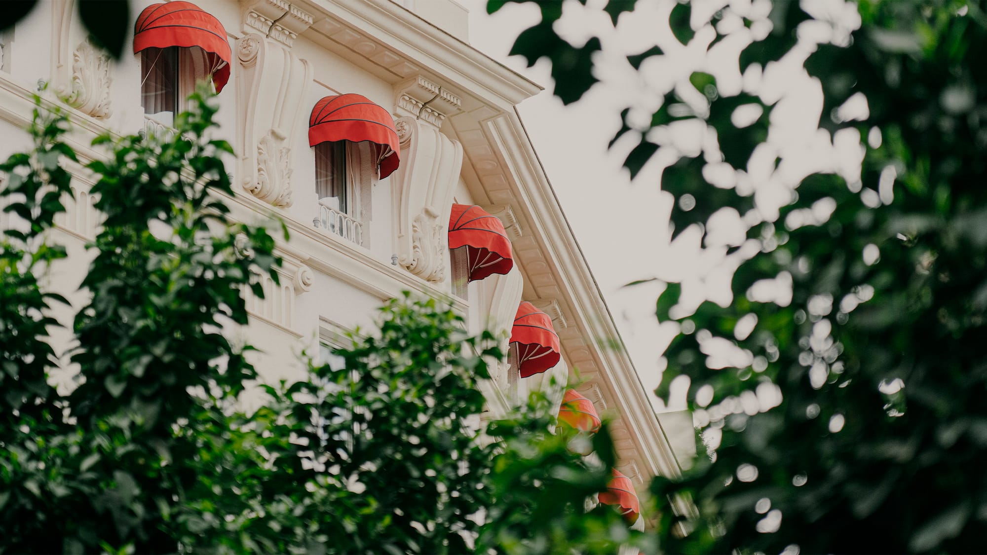 Classic white building facade with red awnings on windows, framed by green trees.