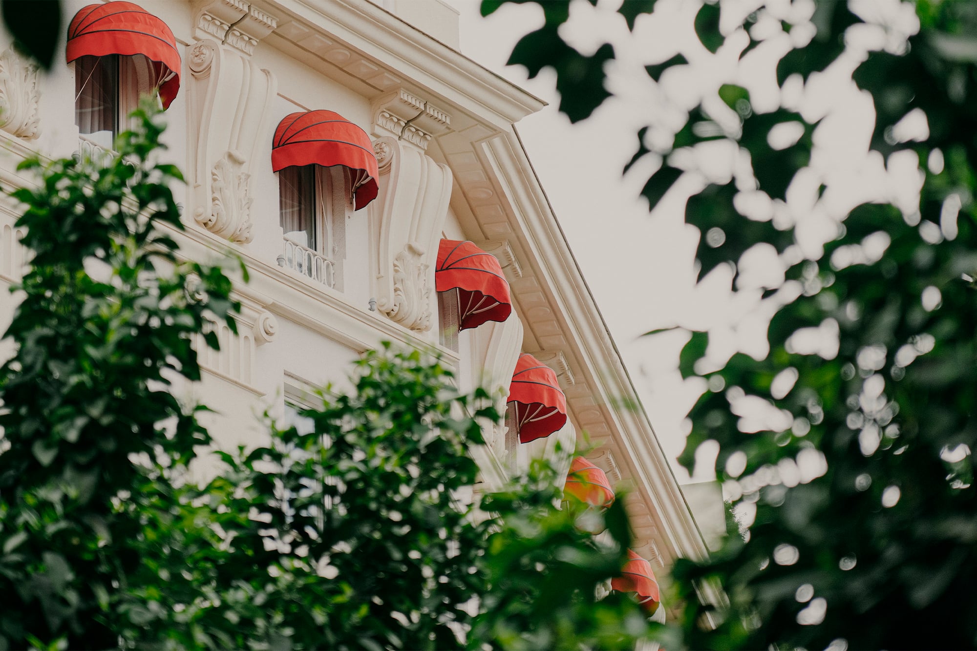 Classic white building facade with red awnings on windows, framed by green trees.
