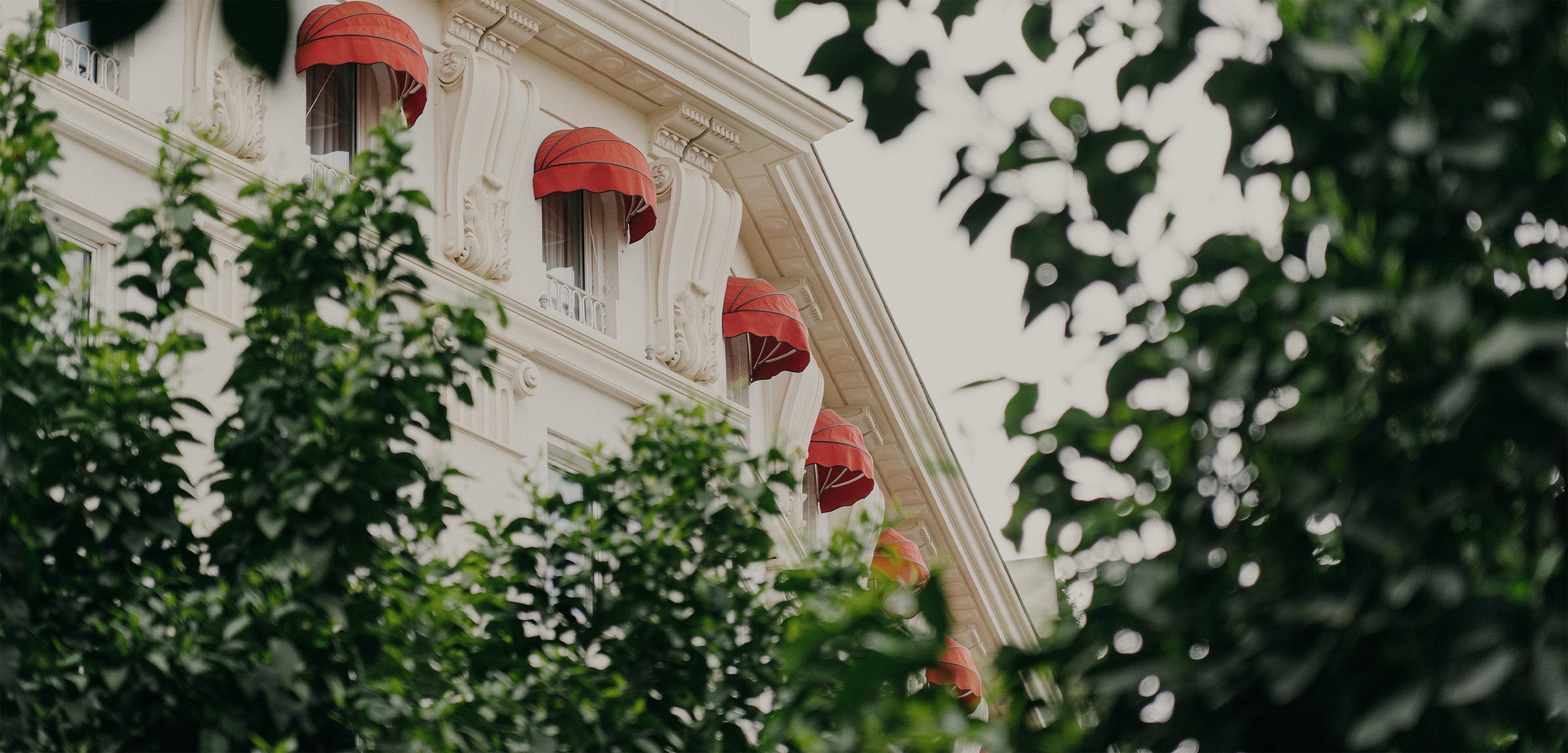 Classic white building facade with red awnings on windows, framed by green trees.