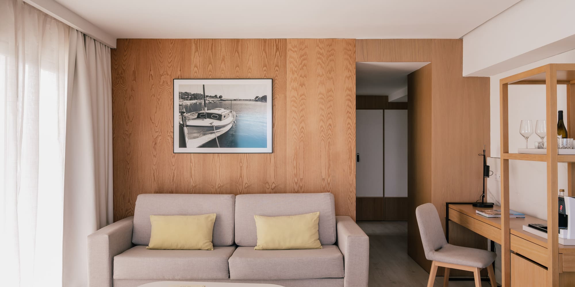 Modern hotel suite living room with sofa, wood wall, desk, and framed boat art.