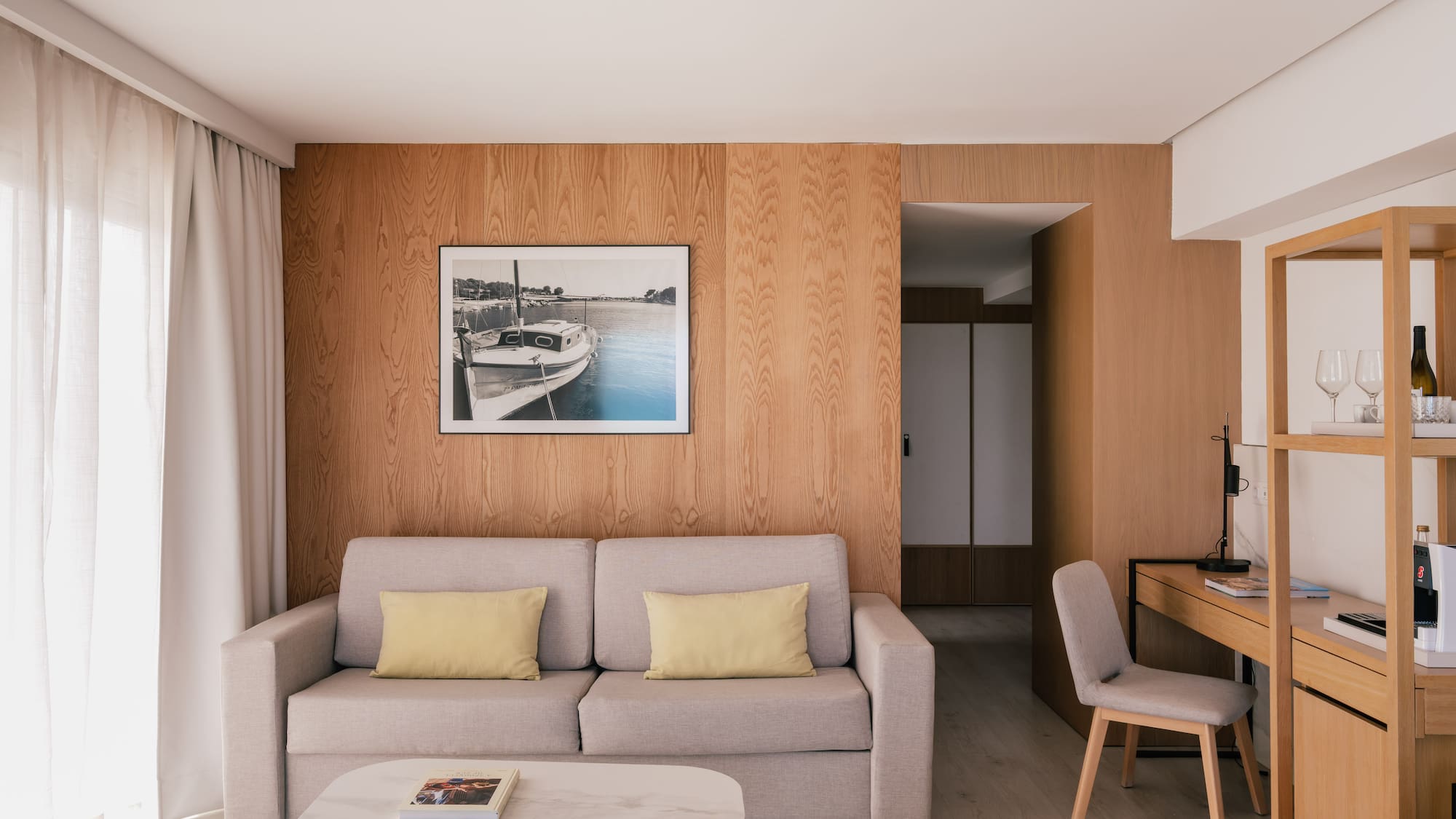 Modern hotel suite living room with sofa, wood wall, desk, and framed boat art.