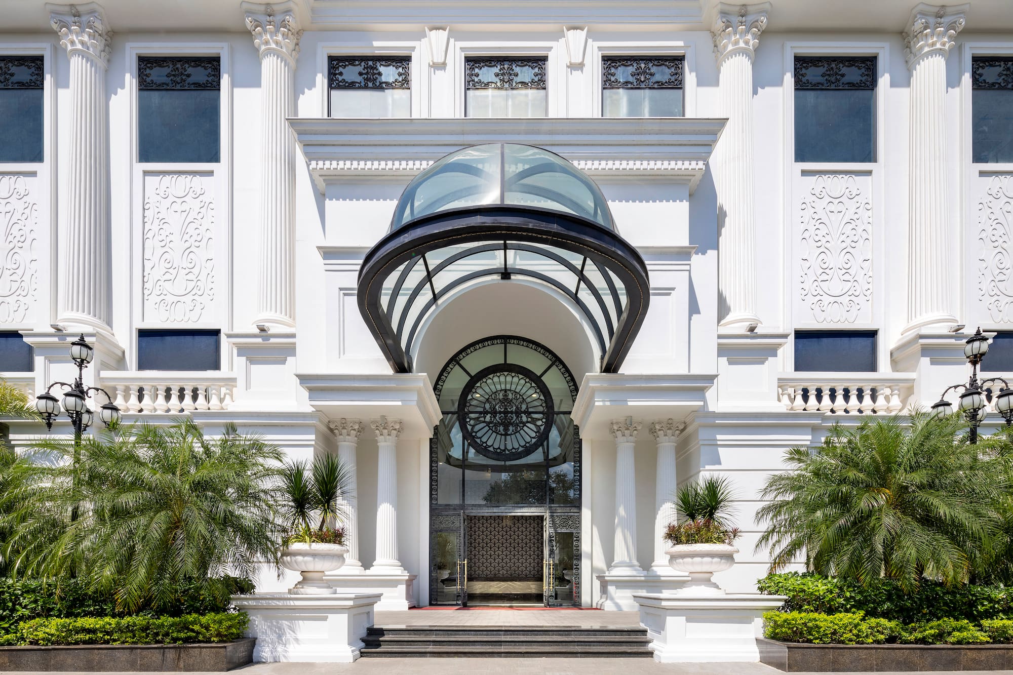 Grand white building entrance with classical columns, glass dome, and palm trees.