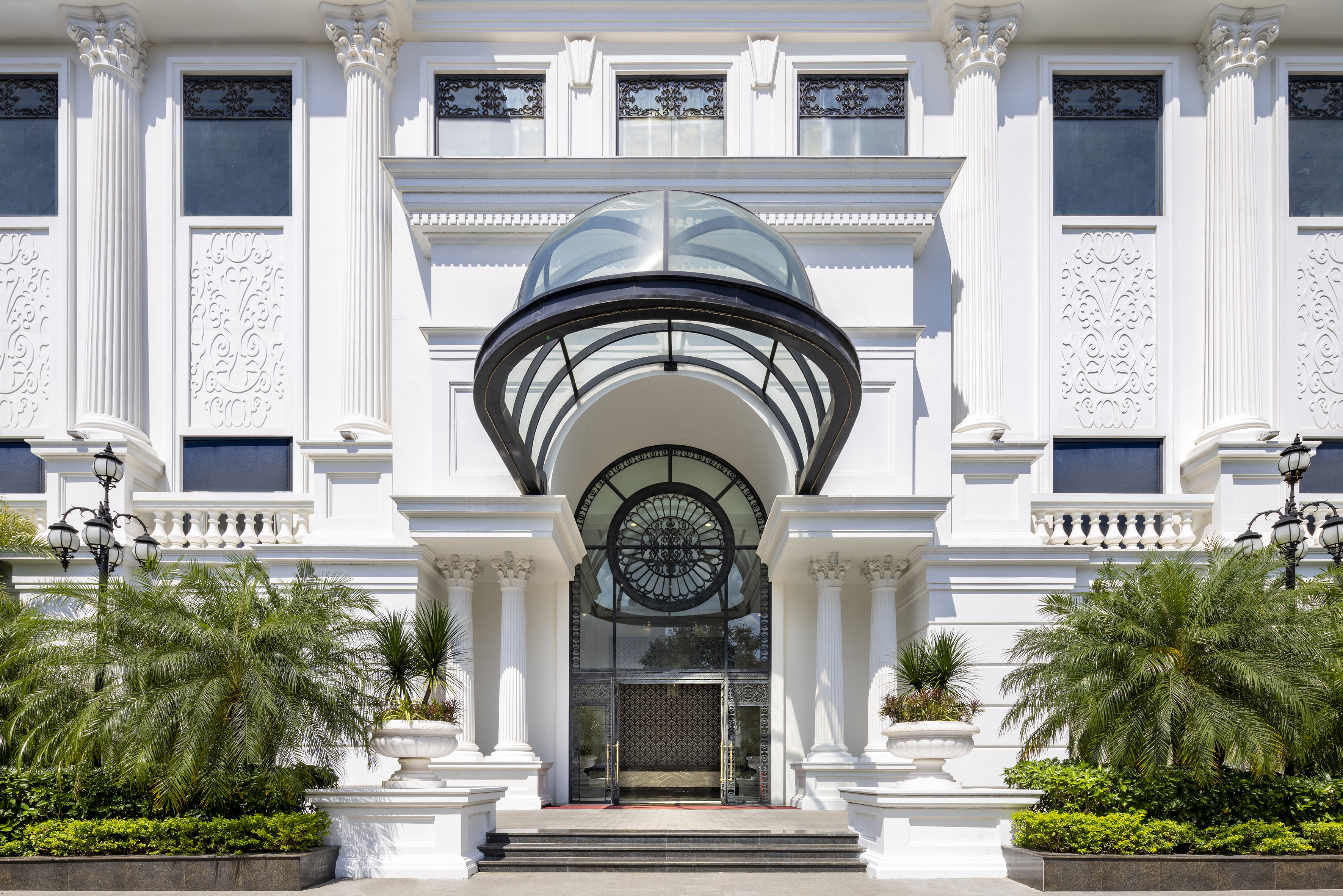 Grand white building entrance with classical columns, glass dome, and palm trees.