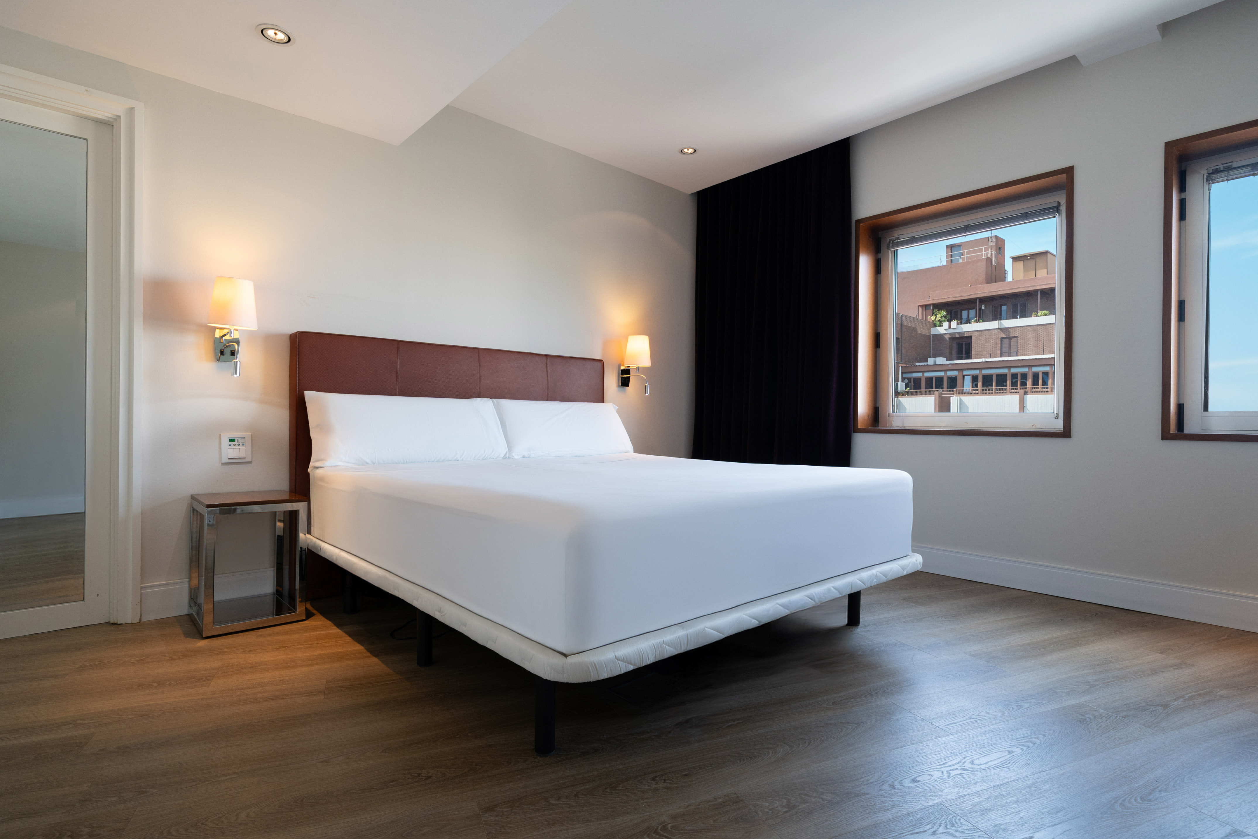 Modern hotel bedroom with king bed, white linen, wood floor, and large windows.