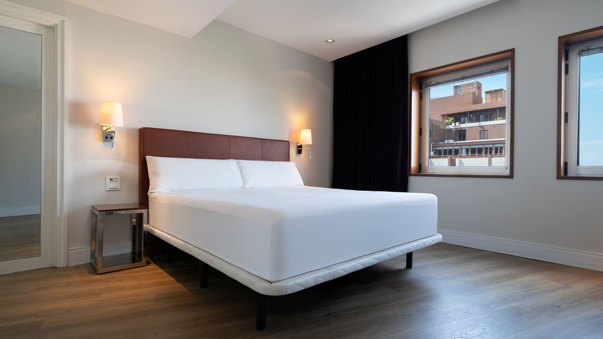 Modern hotel bedroom with king bed, white linen, wood floor, and large windows.