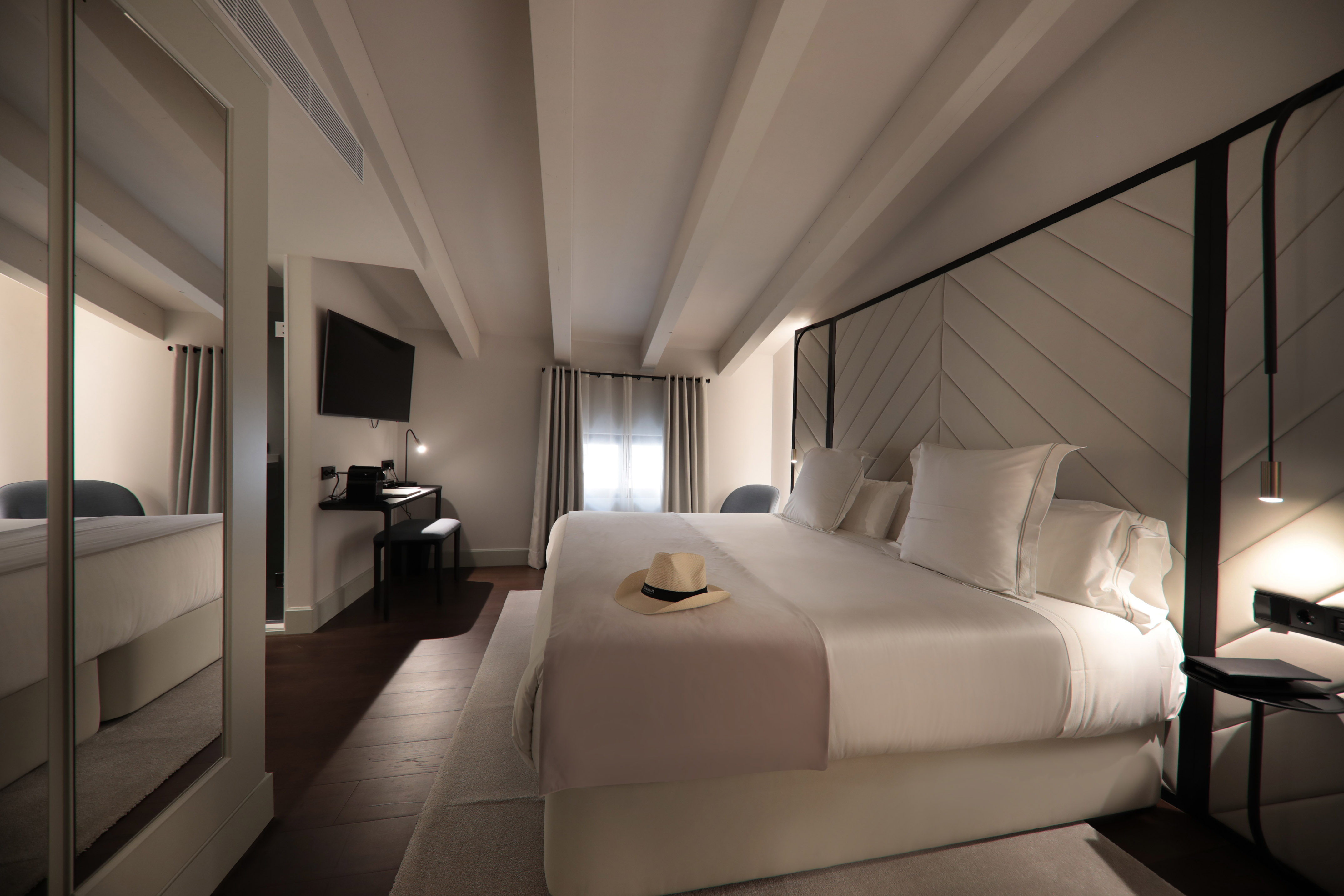 Modern hotel bedroom with king bed, white linen, patterned headboard, and ceiling beams.