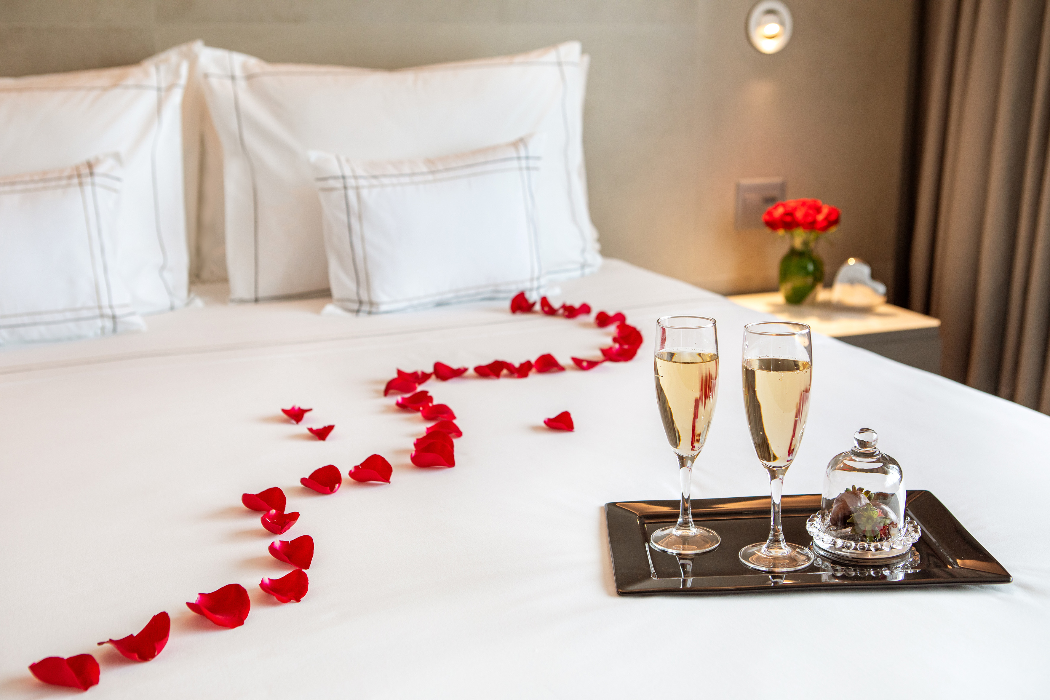 Romantic hotel bed with red rose petals, champagne, and strawberries.