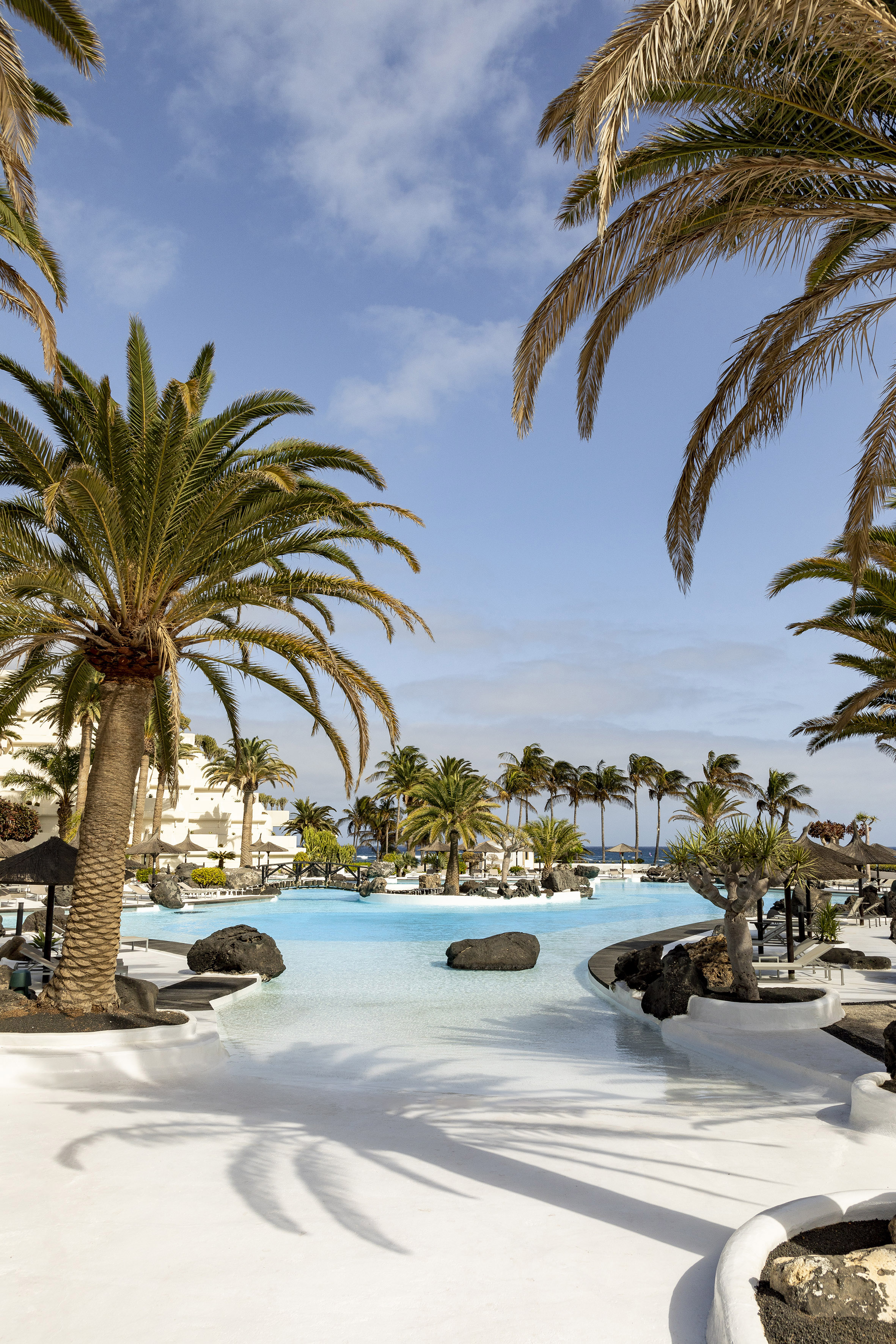 Lanzarote resort pool with palm trees, white buildings, and dark volcanic rocks.