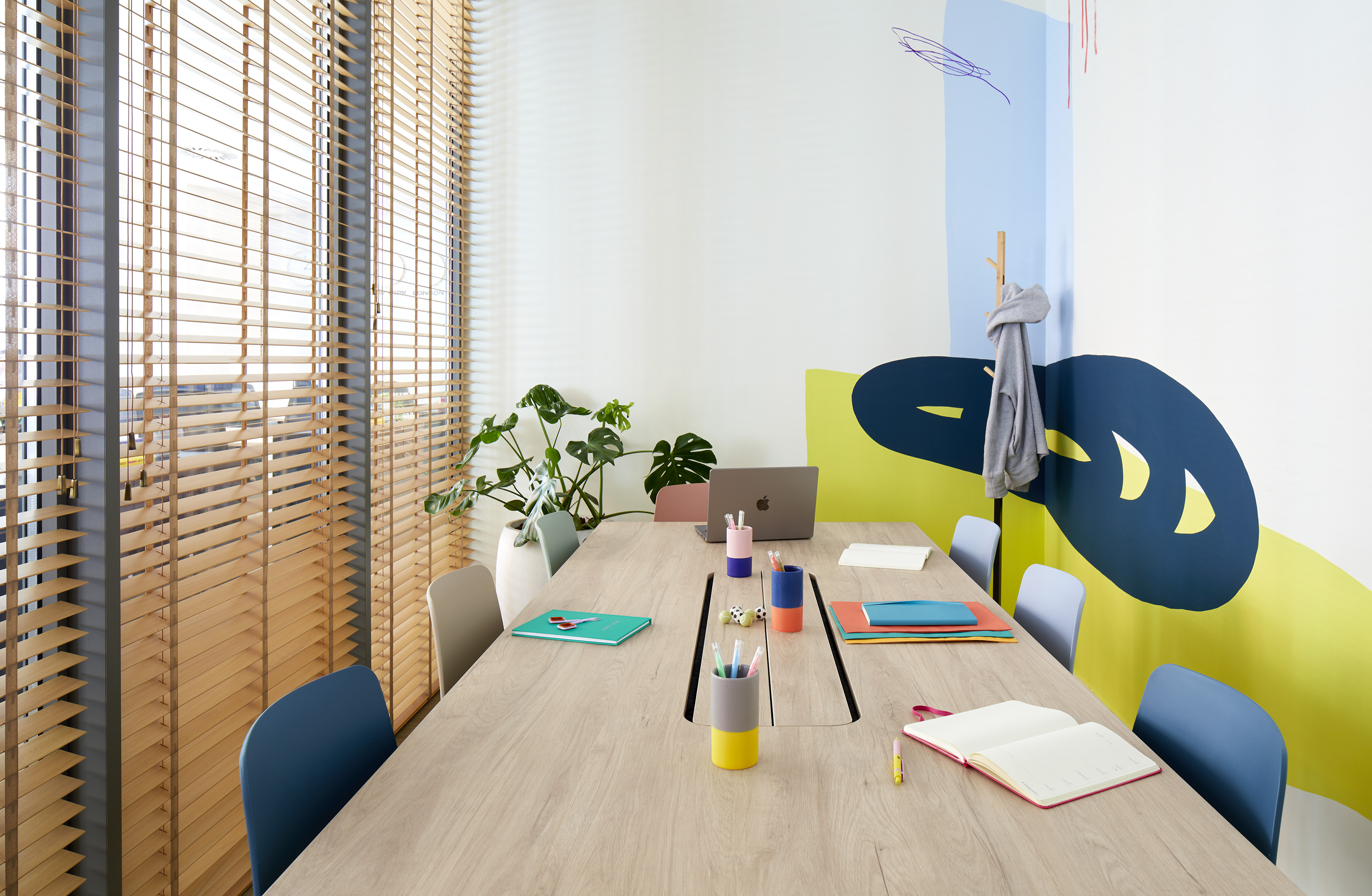 Modern meeting room with long wooden table, colorful chairs, and abstract wall art.