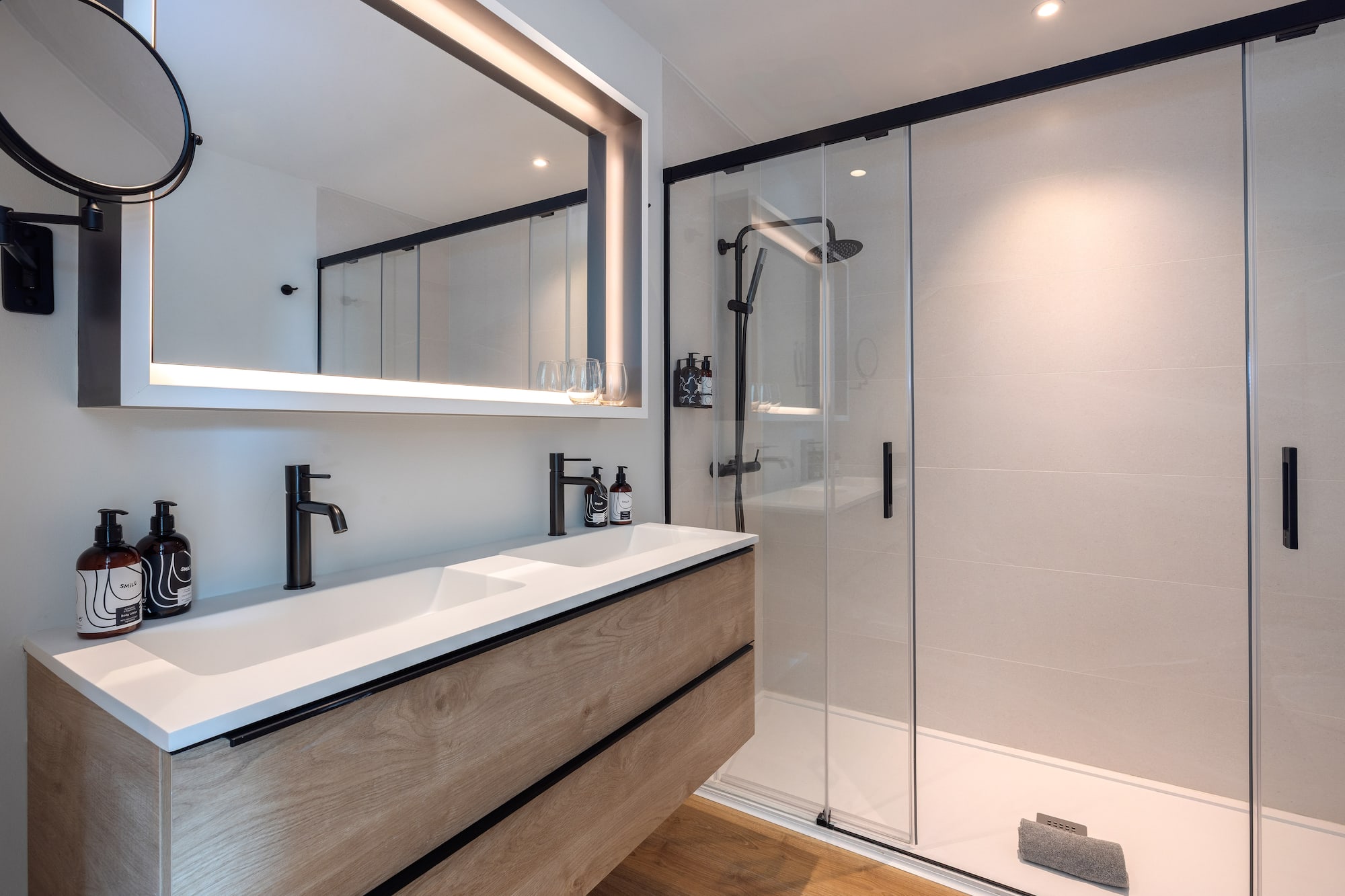 Baño moderno con lavabo de madera, espejo iluminado y ducha de cristal.