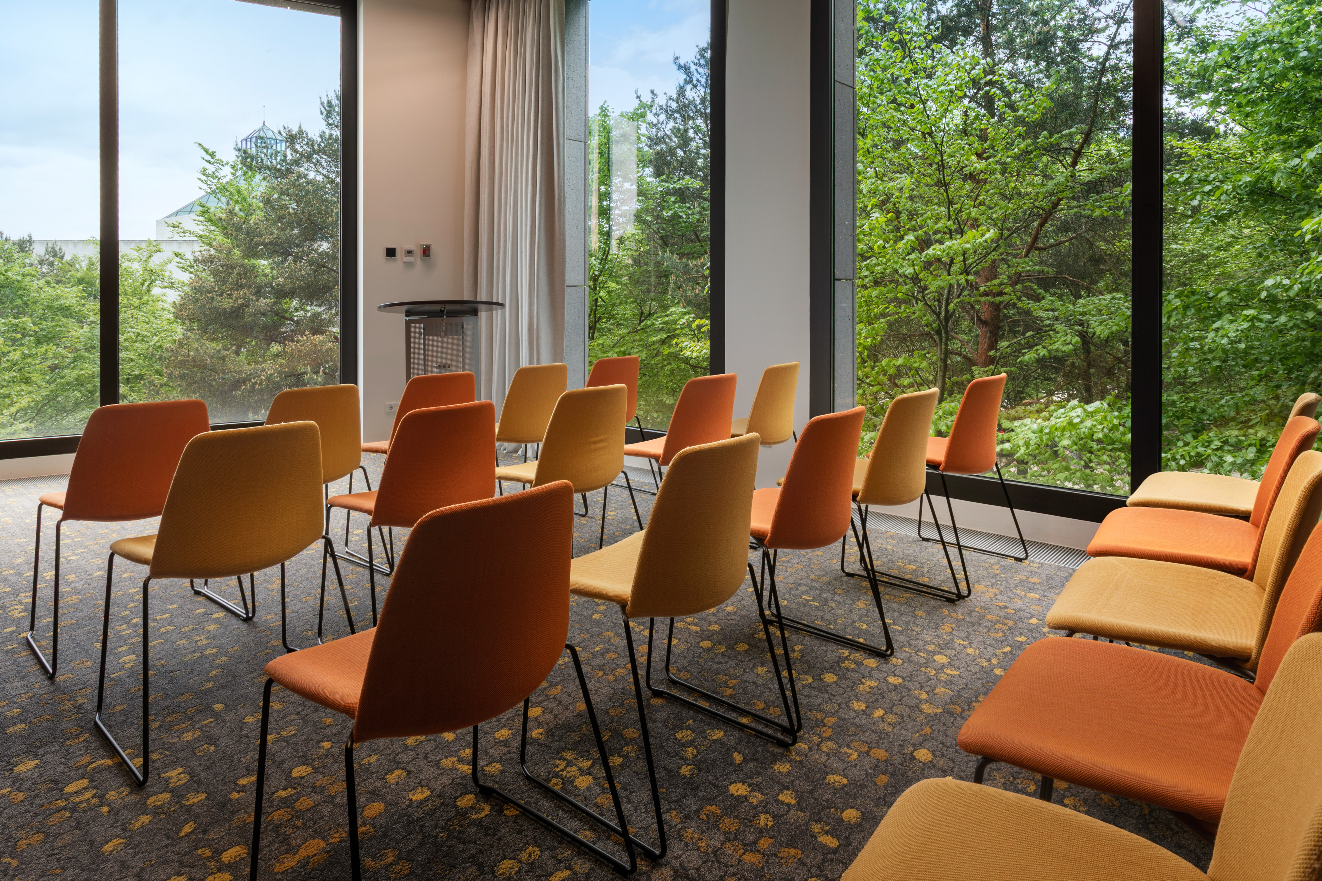 Modern meeting room with orange & yellow chairs, large windows, and forest view.