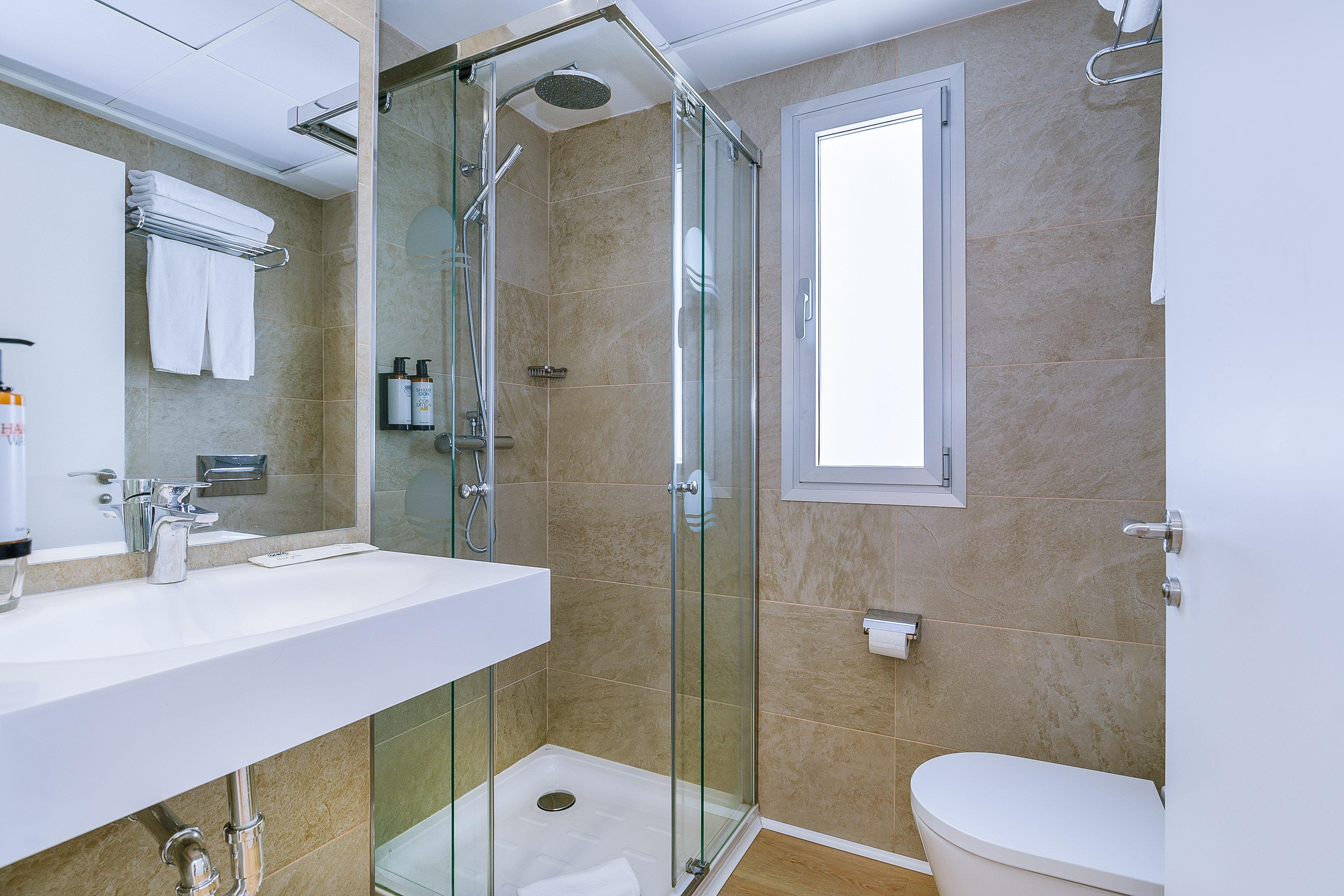 Modern hotel bathroom: glass shower, white sink, toilet, beige tiles.