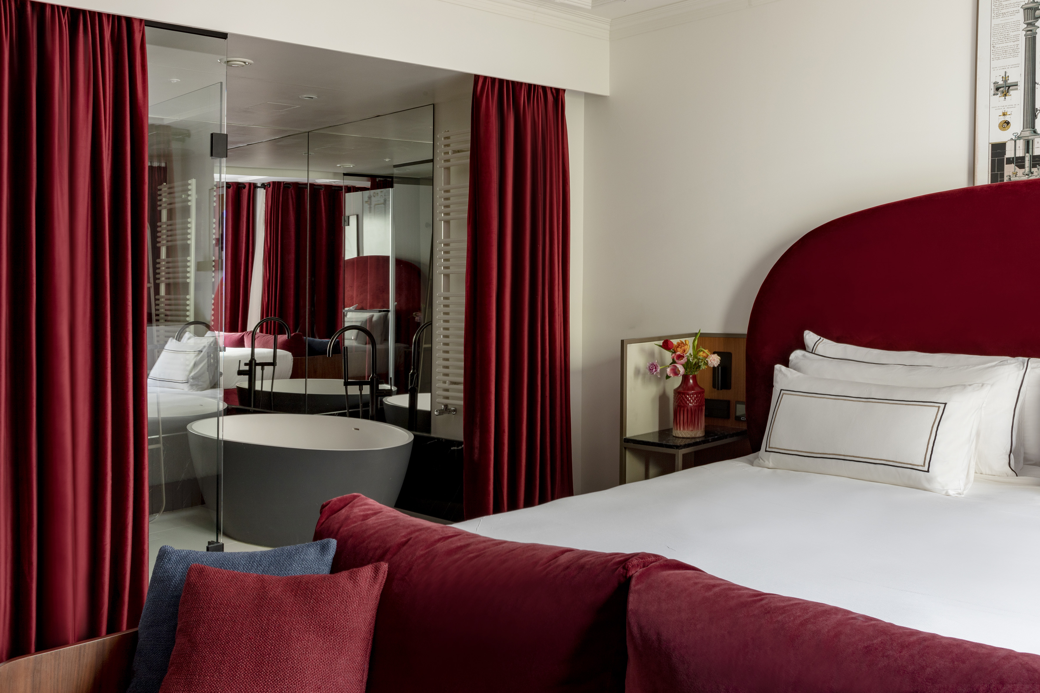 Elegant hotel bedroom with red velvet decor, white bed, and freestanding bathtub.