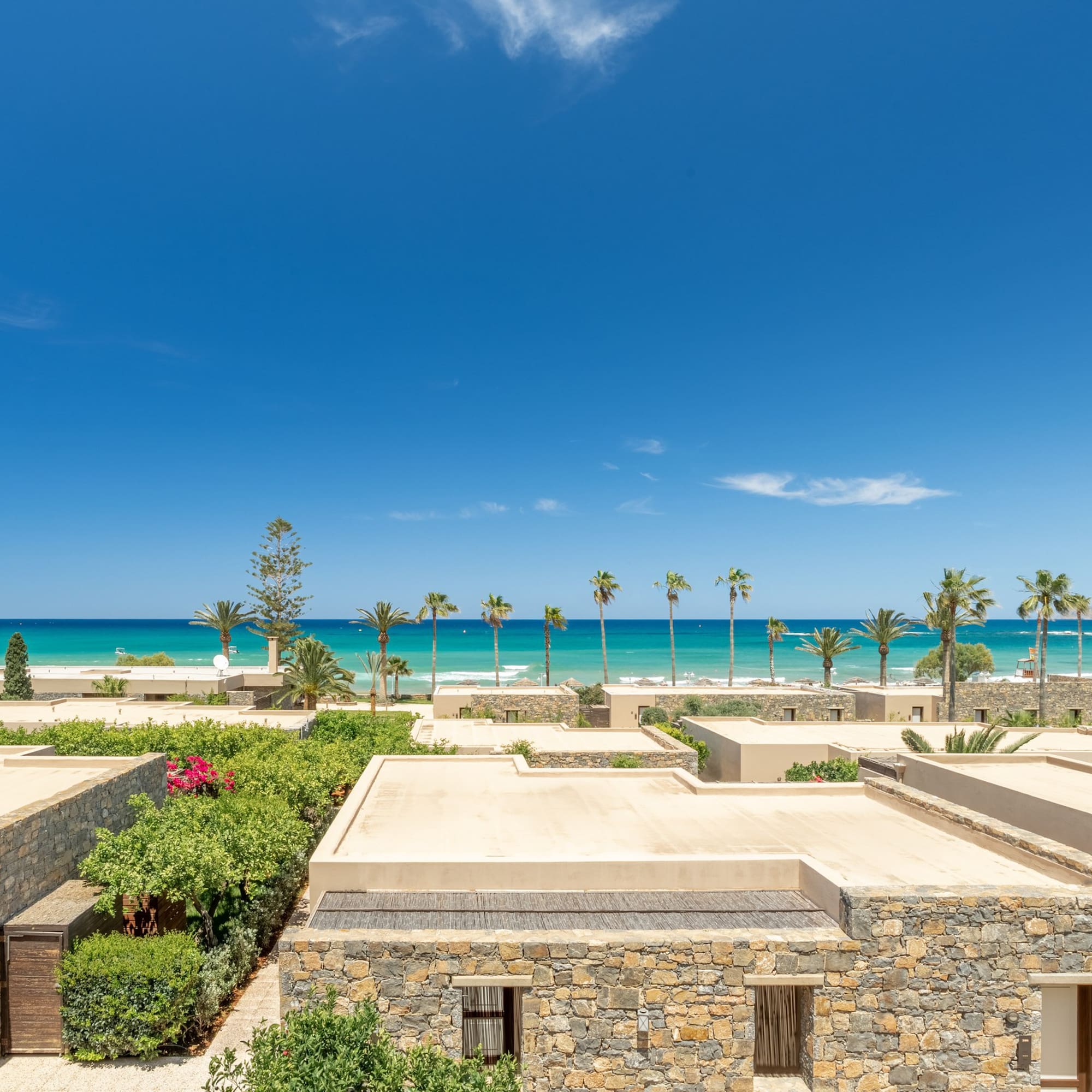 Beachfront stone villas, lush gardens, turquoise sea, and palm trees.