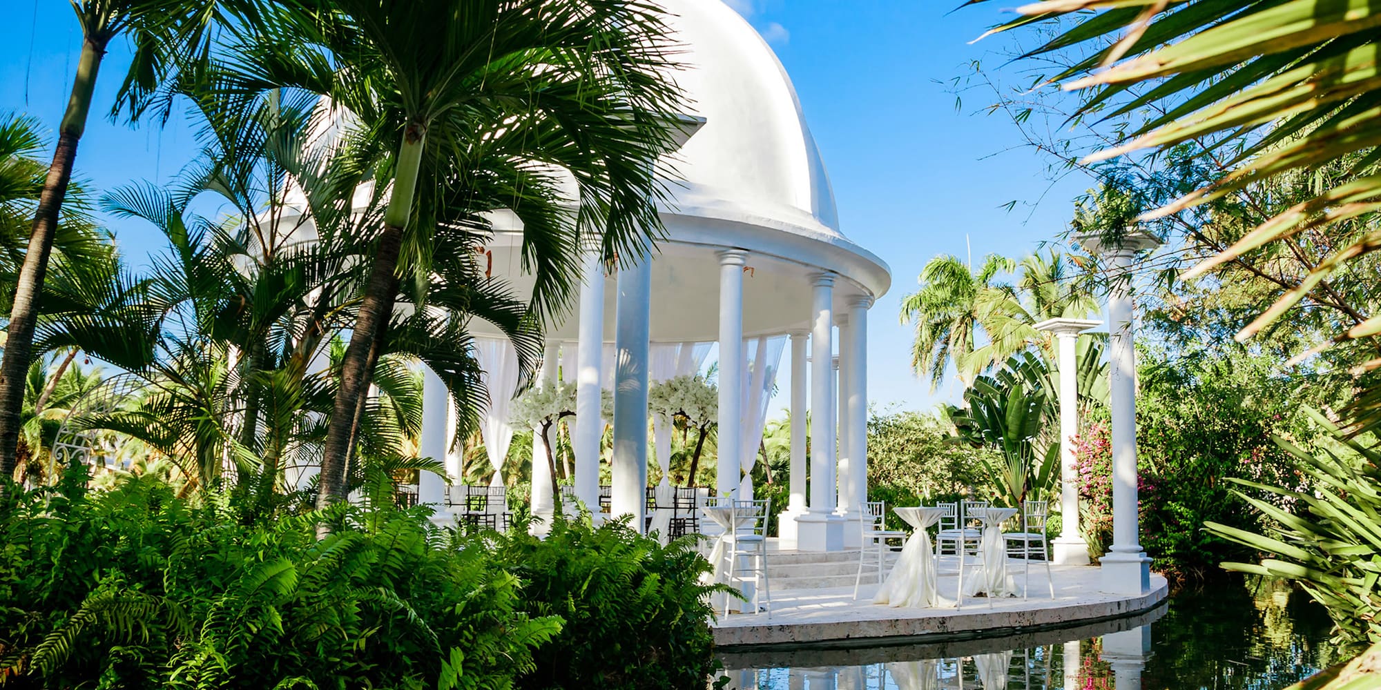 Tropical wedding gazebo with white dome, palm trees, and water reflection.