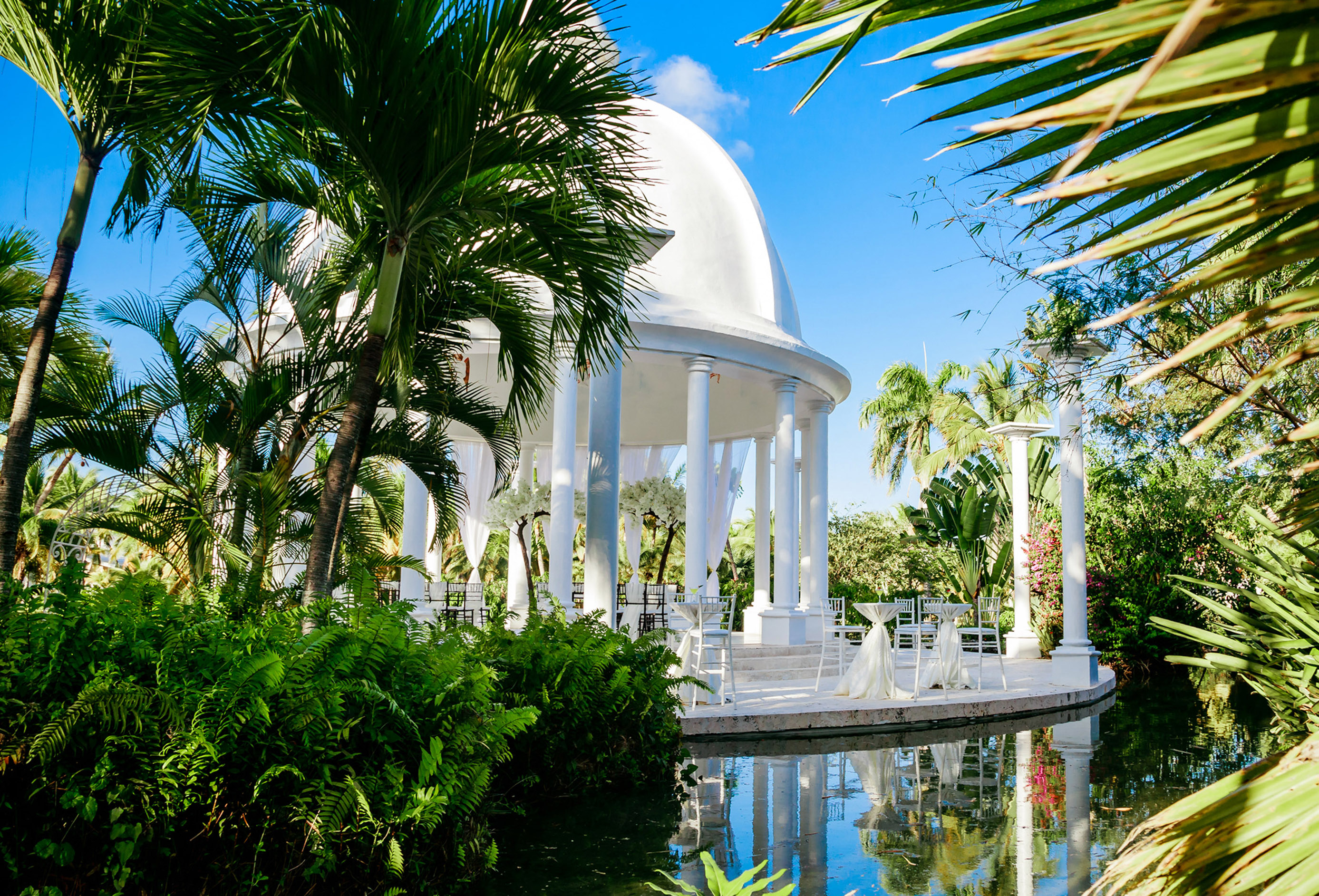 Tropical wedding gazebo with white dome, palm trees, and water reflection.