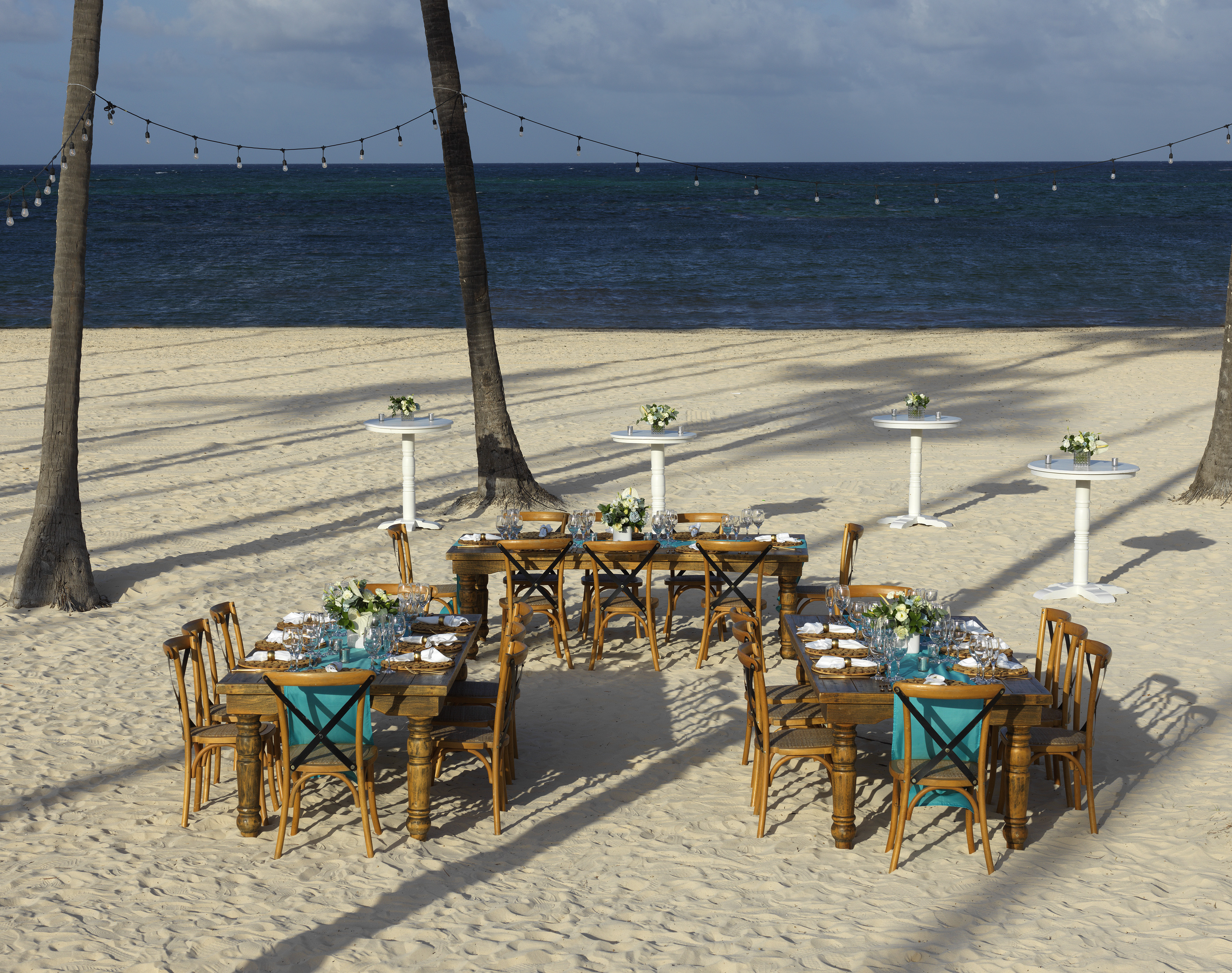 Elegant beach dining setup with wooden tables, teal runners, palm trees, ocean.